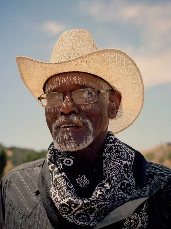 An elderly man wearing a large straw cowboy hat, glasses, and a black bandana with white paisley patterns, outdoors with a clear sky in the background.