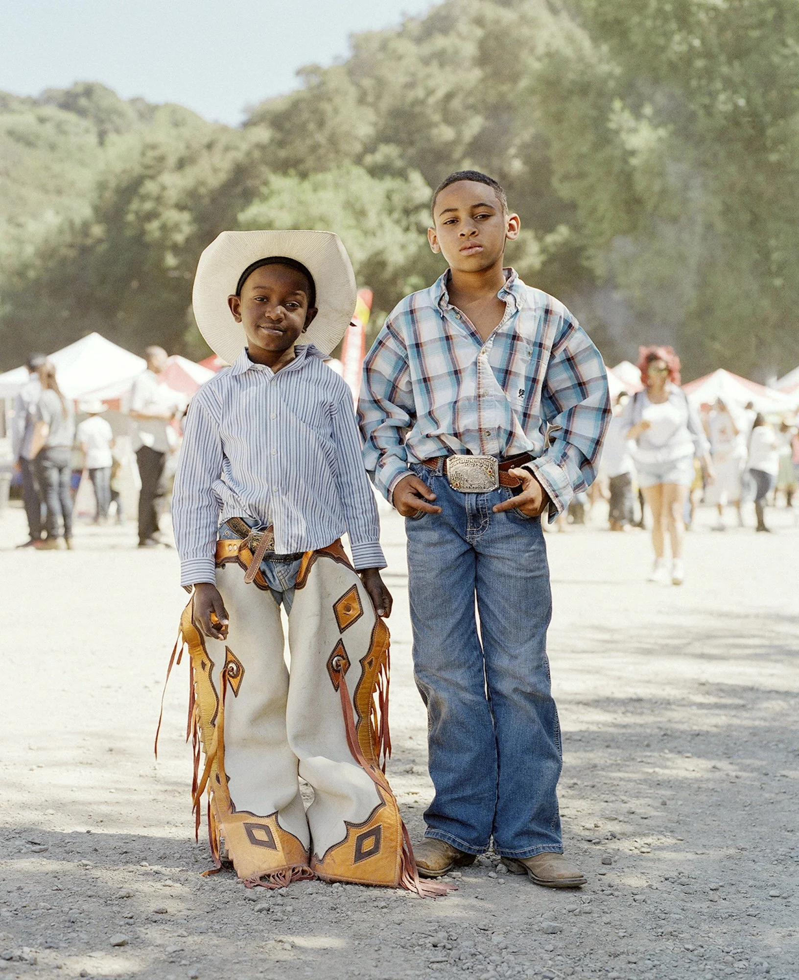 Two young boys dressed in cowboy attire standing outdoors at an event or fair, with tents and other people in the background.