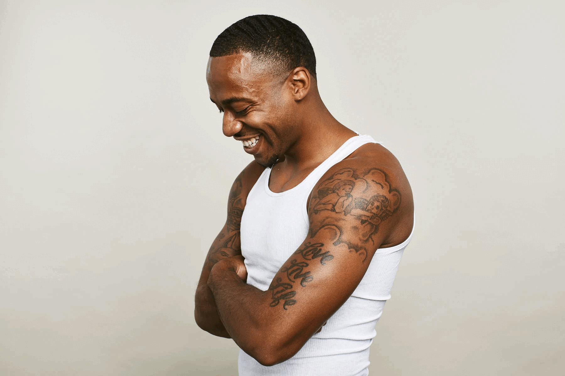 A young man smiling and looking down, wearing a white tank top. He has tattoos on his arms and is standing against a plain, light-colored background.