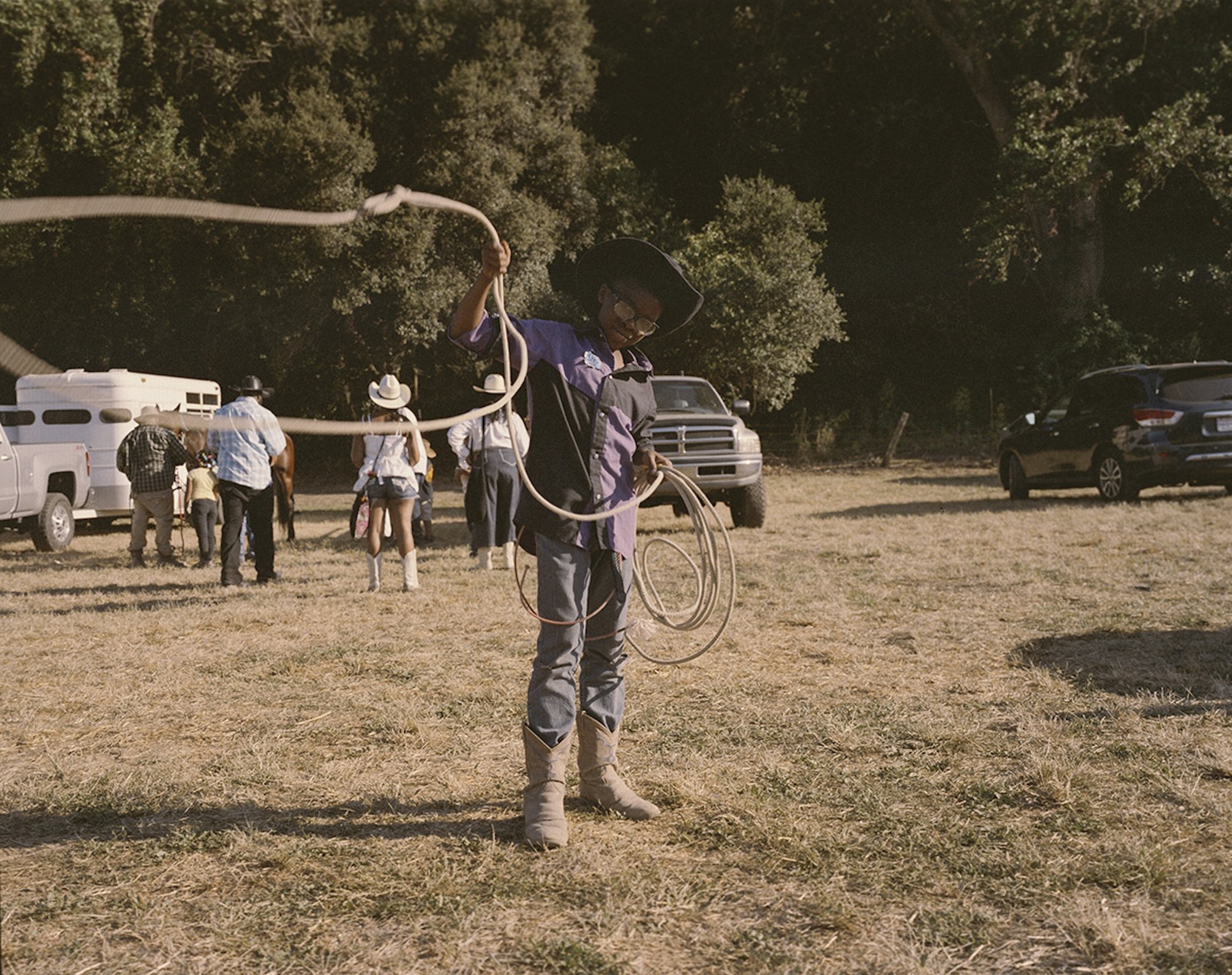 A person in cowboy hat and boots holding a lasso in a field with other people and cars in the background, some people wearing cowboy hats.