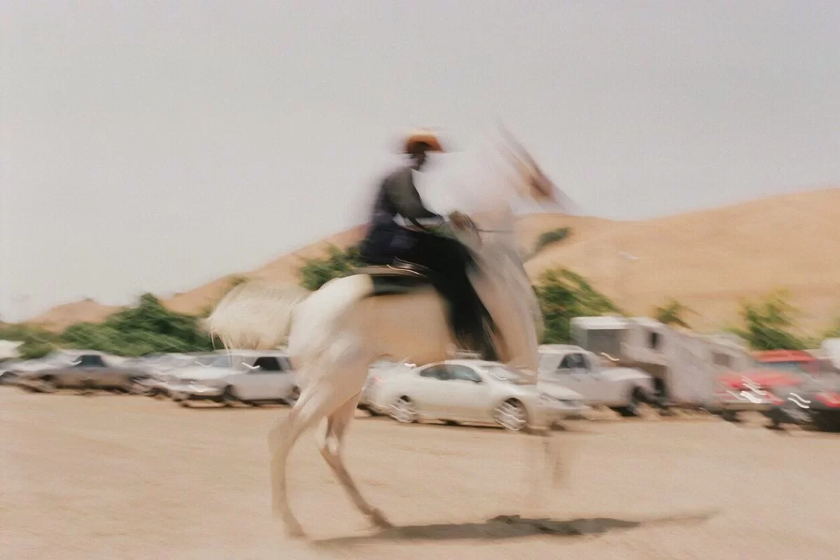 A person riding a camel in a desert area with parked cars in the background.