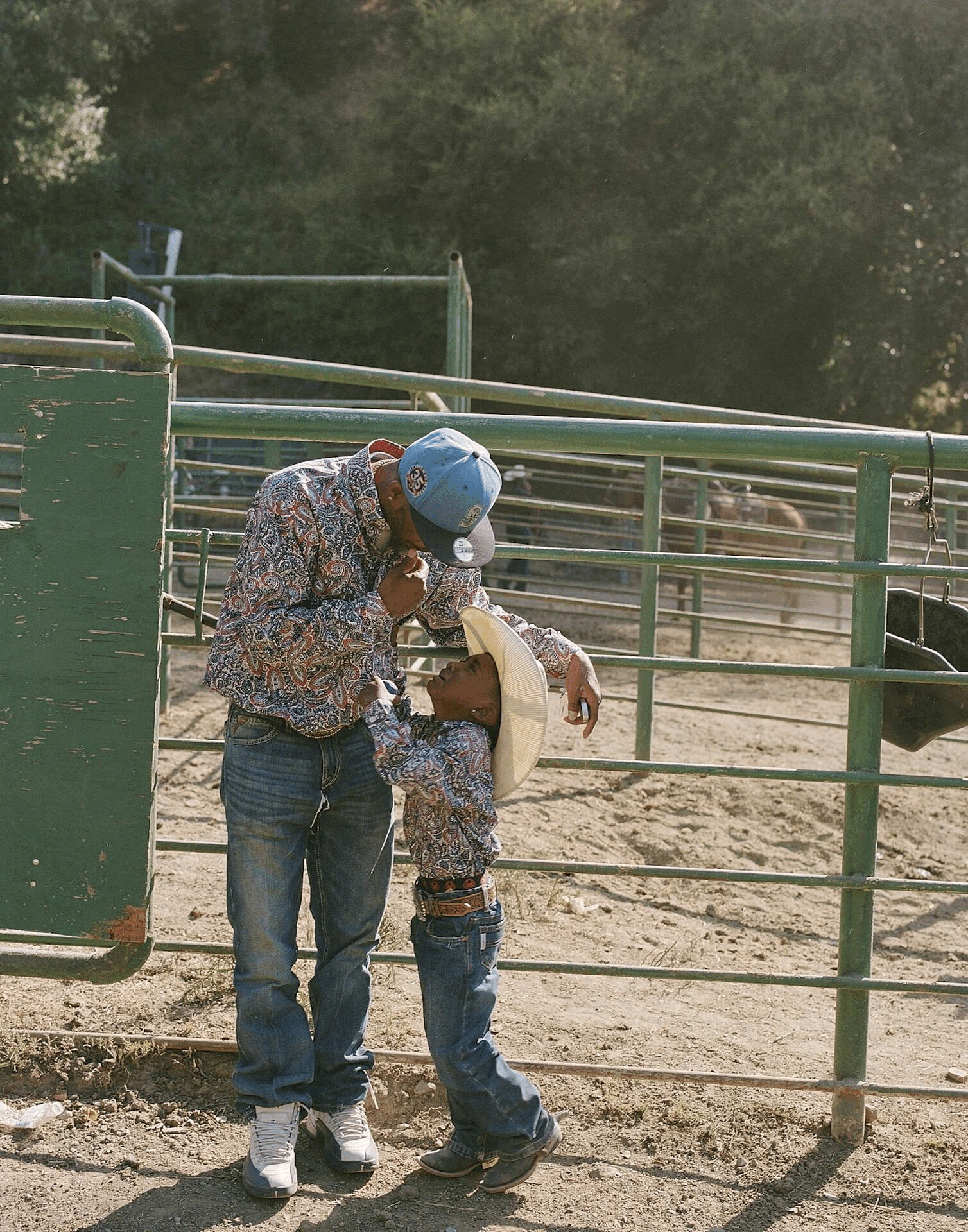 A man and a young boy in cowboy hats and western-style clothing exchanging a kiss on a farm or ranch, with metal fencing and a dirt ground in the background.