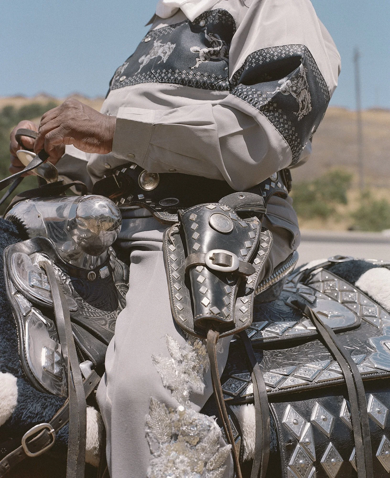 Close-up of a person wearing a western shirt, sitting on a horse with silver and black decorative tack, including a bridles and saddle, with a desert landscape background.