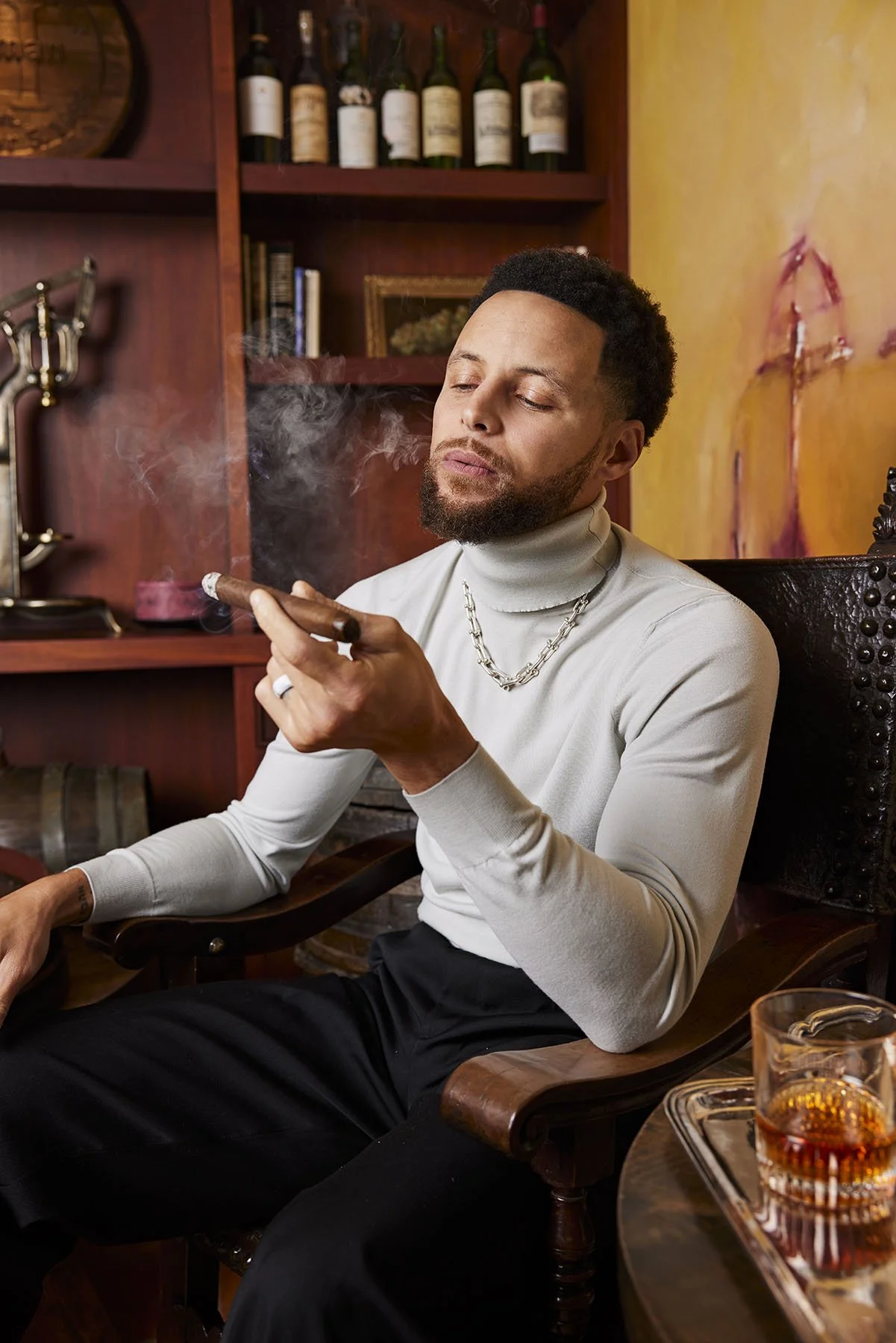 A man sitting in a wooden armchair, smoking a cigar, in a cozy room with wooden shelves, wine bottles, and a glass of liquor on a tray.
