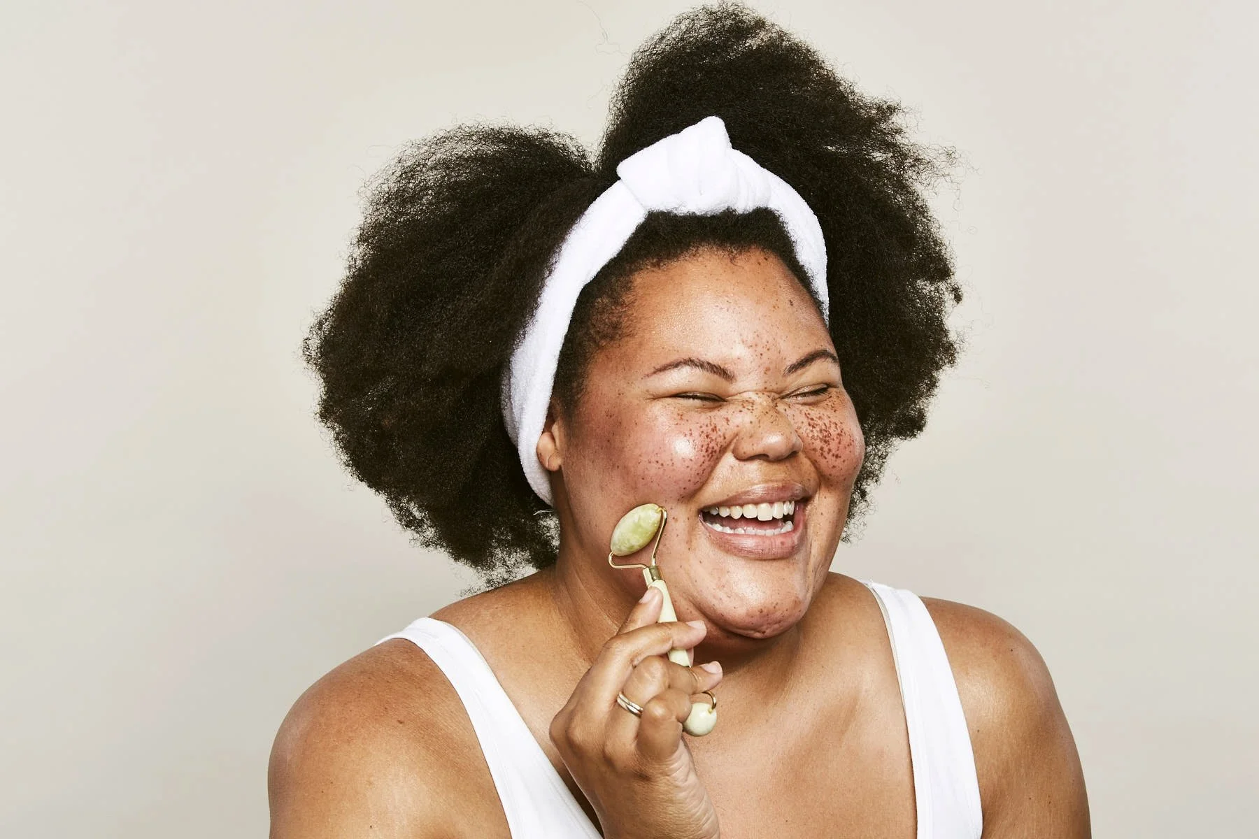 A woman with curly hair, wearing a white headband, smiling and holding a jade roller to her cheek, in front of a plain white background.