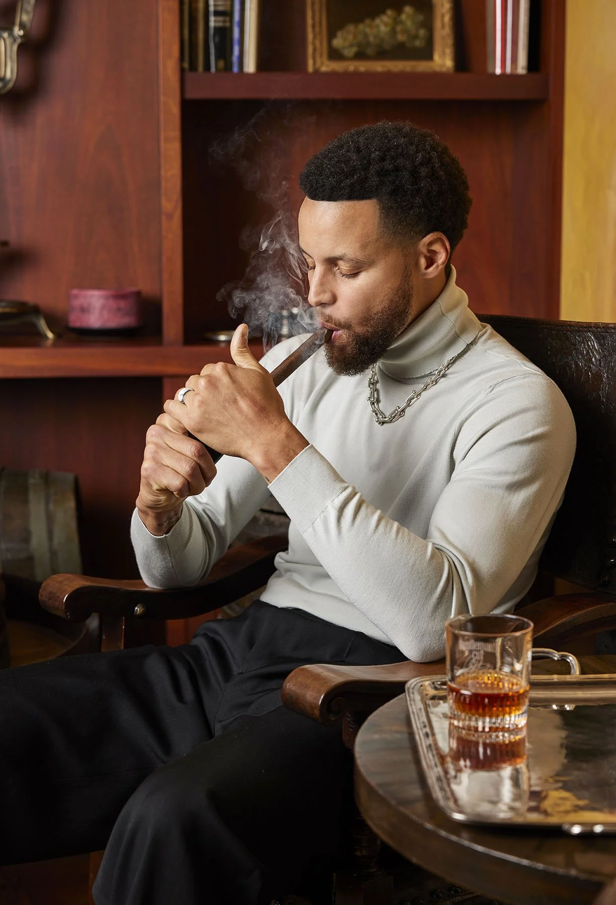 A man with dark curly hair and a beard, wearing a light-colored turtleneck sweater and a chain necklace, sitting in a wooden chair, smoking a cigar in a room with wooden shelves and a framed picture in the background. He has a glass of whiskey on a tray on the table next to him.