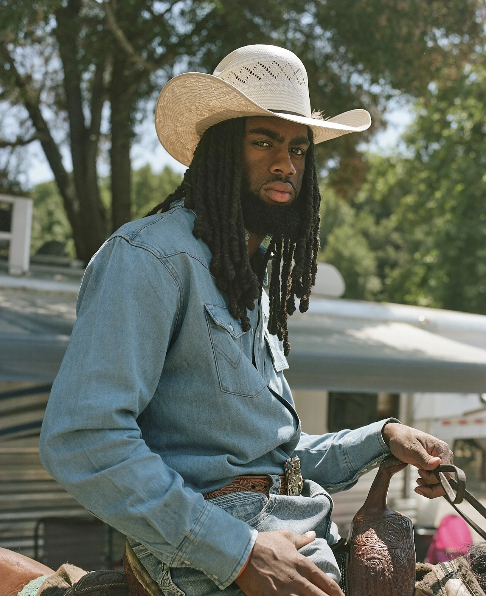 A man wearing a white cowboy hat, a denim jacket, and jeans at an outdoor event with trees in the background.