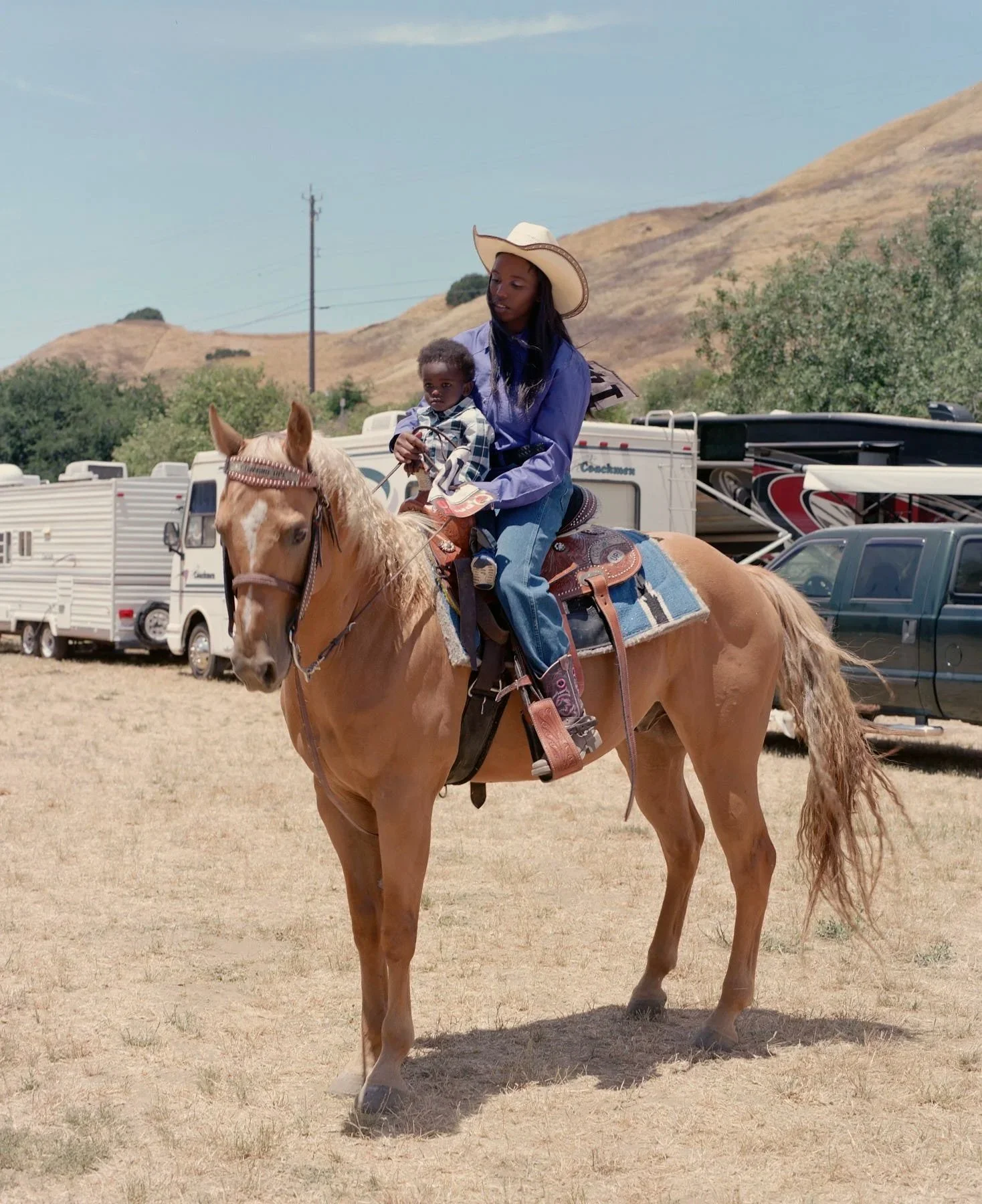 A woman riding a horse with a young child on her lap in an outdoor setting, with trailers and trucks in the background and hills in the distance.