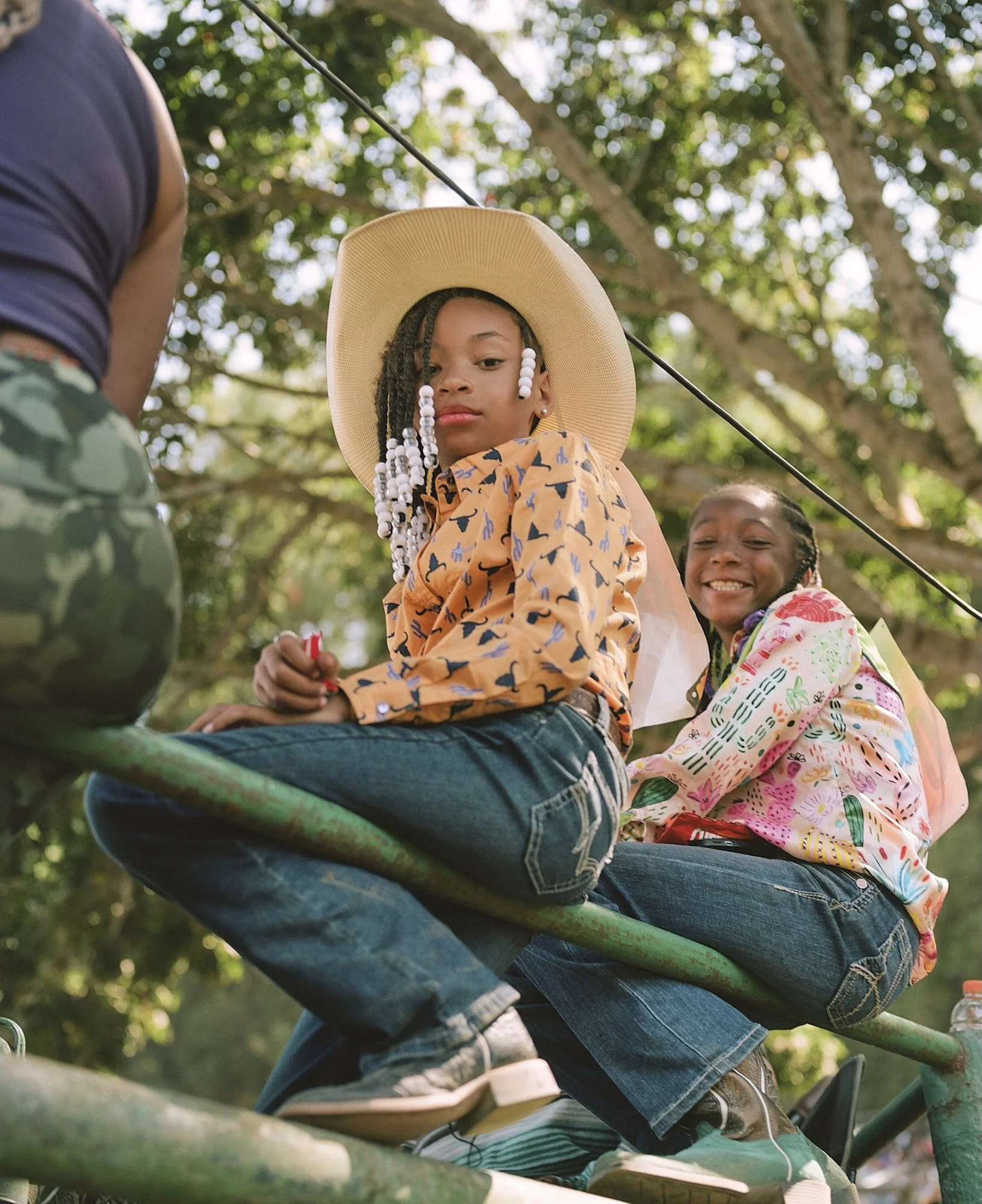 Two young girls sitting on a green park bench, smiling, with trees in the background. One girl is wearing a large sun hat and has beads in her hair, while the other girl is smiling widely and wearing a colorful floral jacket.