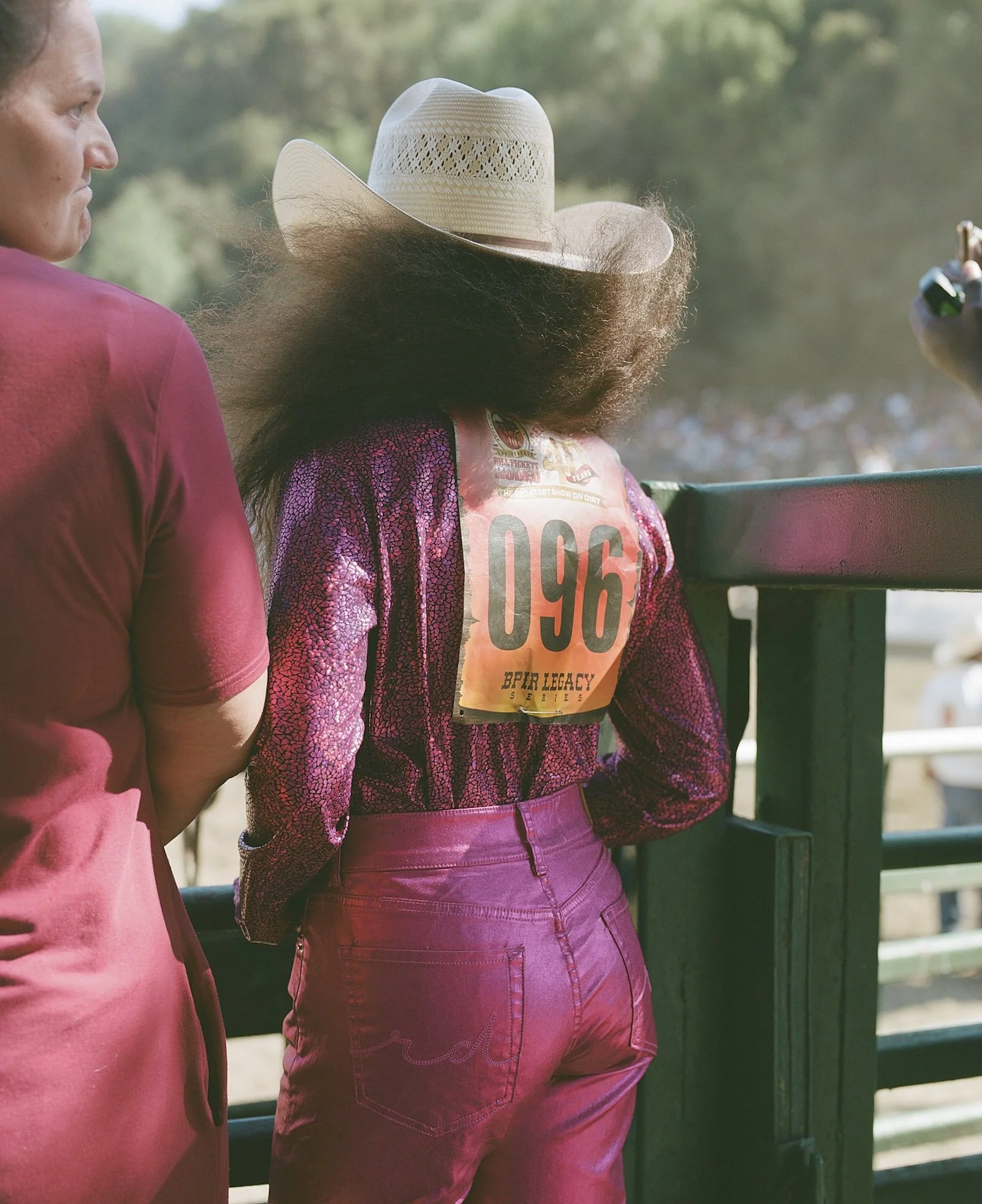 A woman with long curly hair wearing a cowgirl hat and pink cowgirl outfit, standing at a rodeo event, viewed from behind, with a race number '096' on her back.