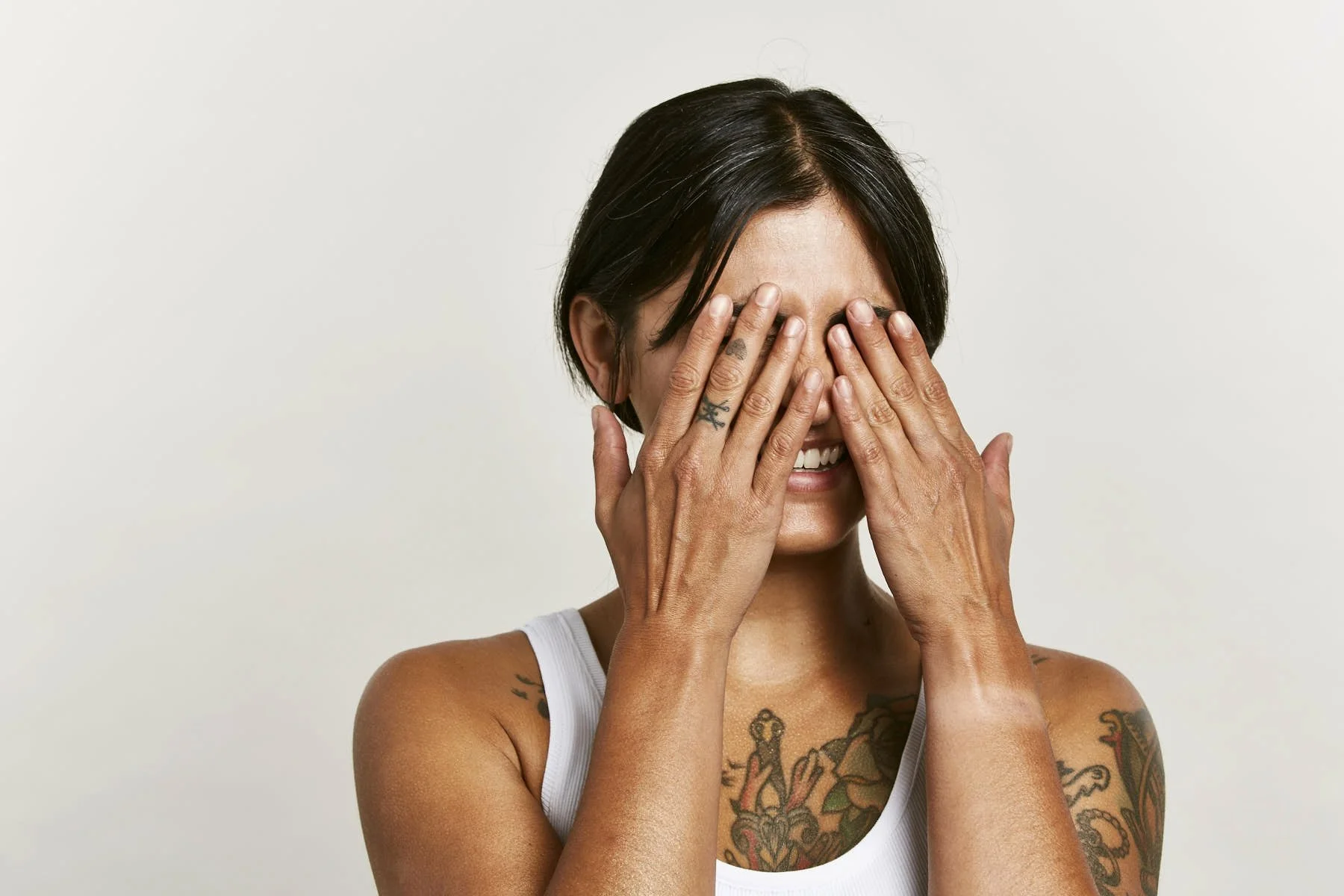 A woman with short dark hair and tattoos covering her arms and chest, holding her hands over her face and smiling.