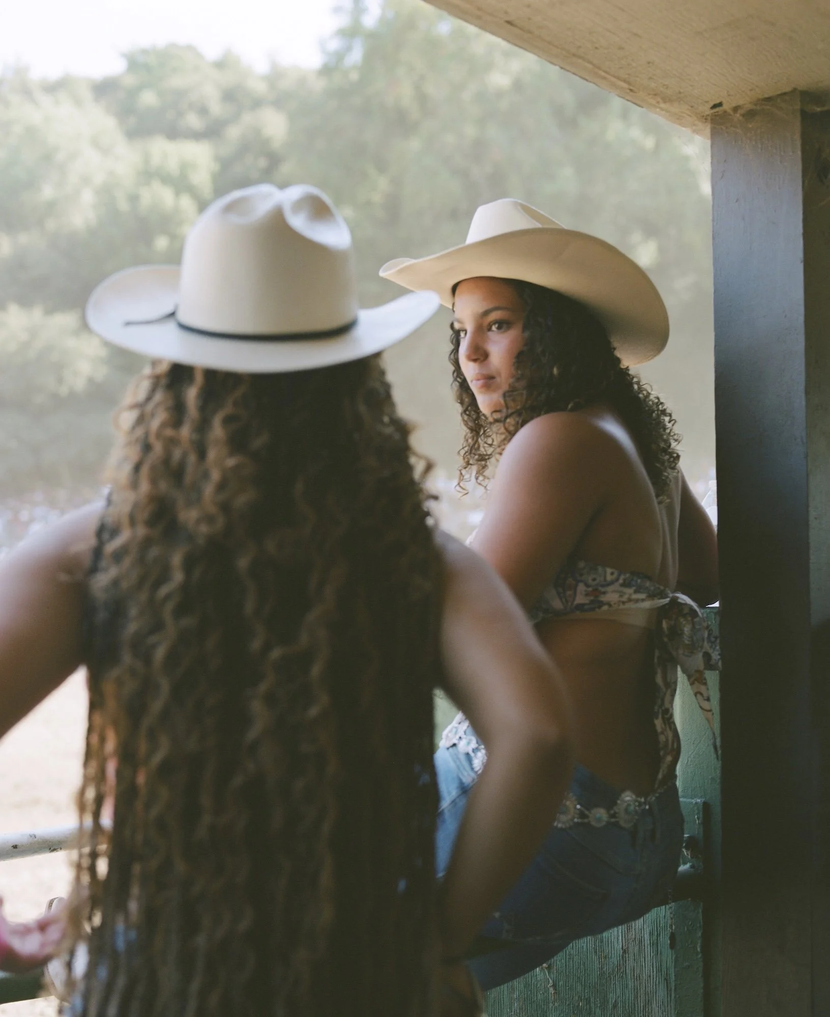 Two women wearing cowboy hats and sleeveless tops, sitting and talking outdoors, with a blurred natural background.