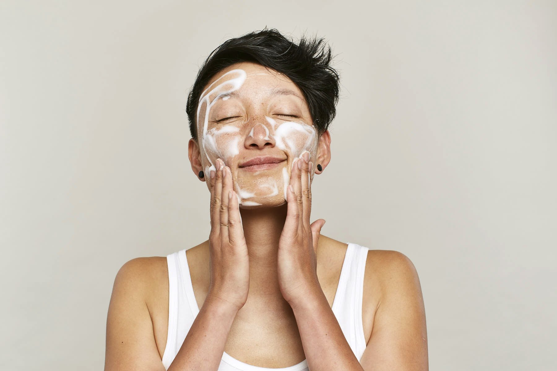 Commercial beauty image of a woman applying face moisturizer with closed eyes, smiling, against plain wall background.