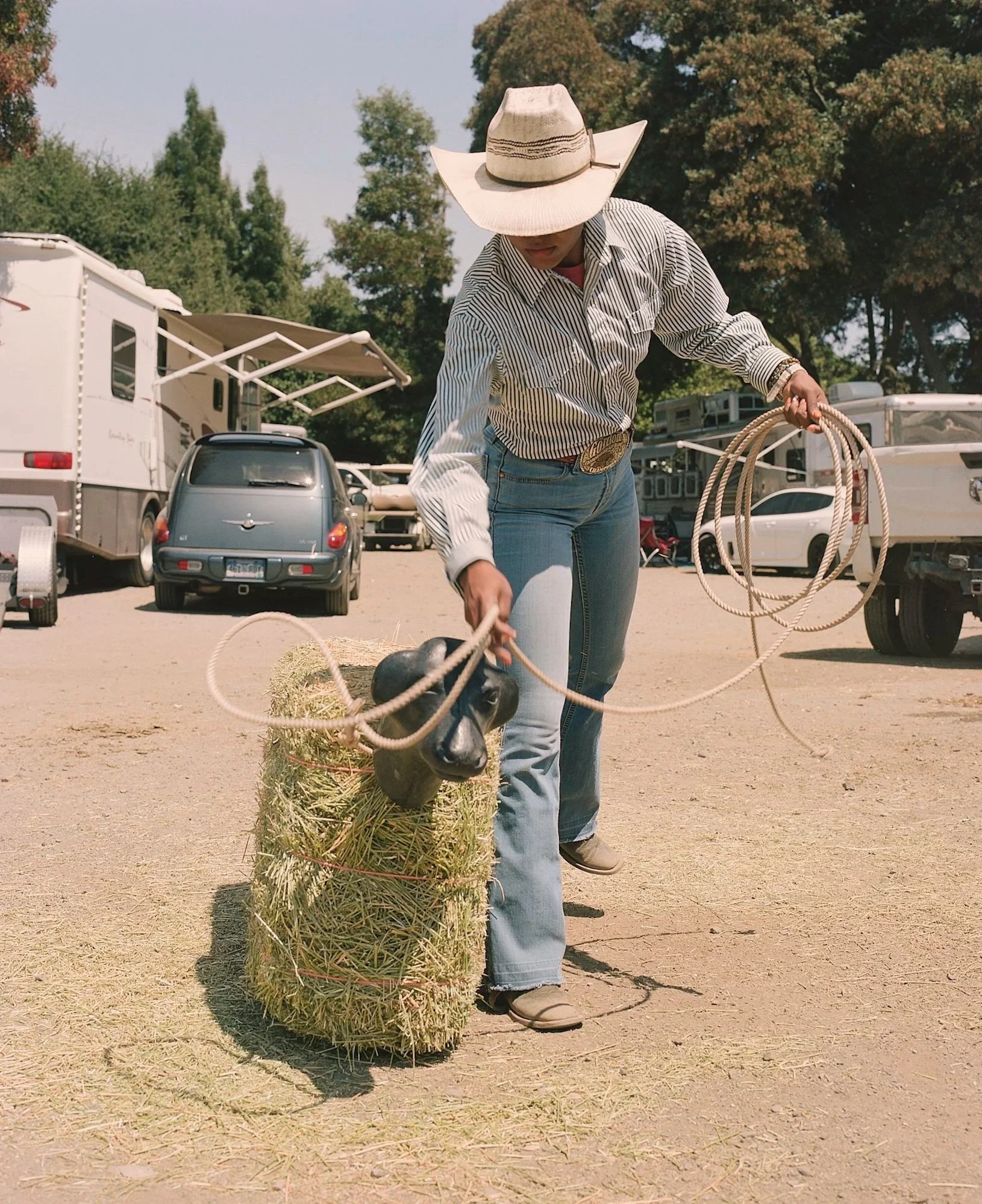 A woman wearing a cowboy hat, striped shirt, and jeans is securing a hay bale to a small horse with a black head. She is holding a rope, and there are parked RVs and cars in the background on a sunny day.