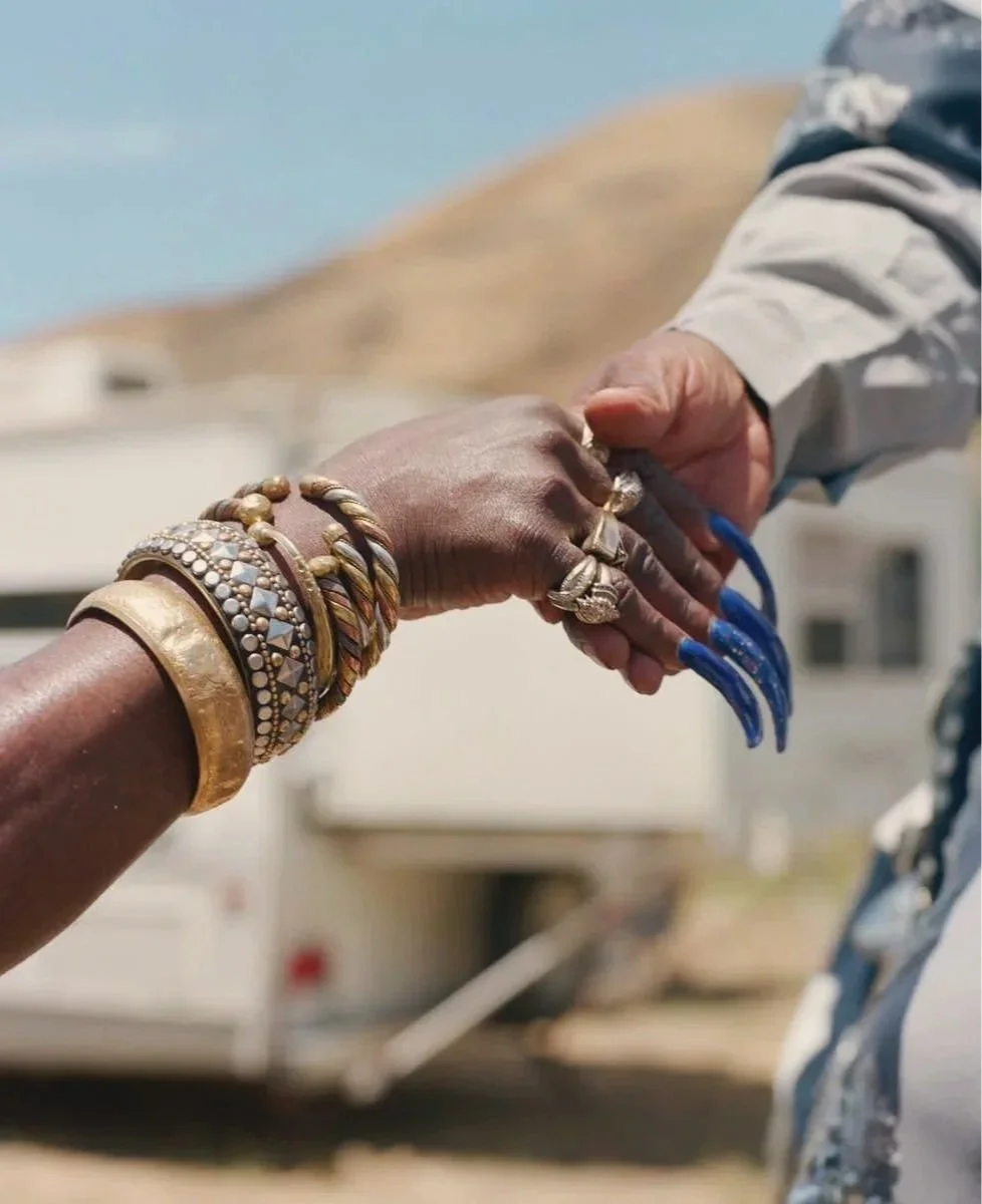 Two people shaking hands, one with multiple bracelets and rings, and the other with long blue acrylic nails, outdoors with a trailer in the background.