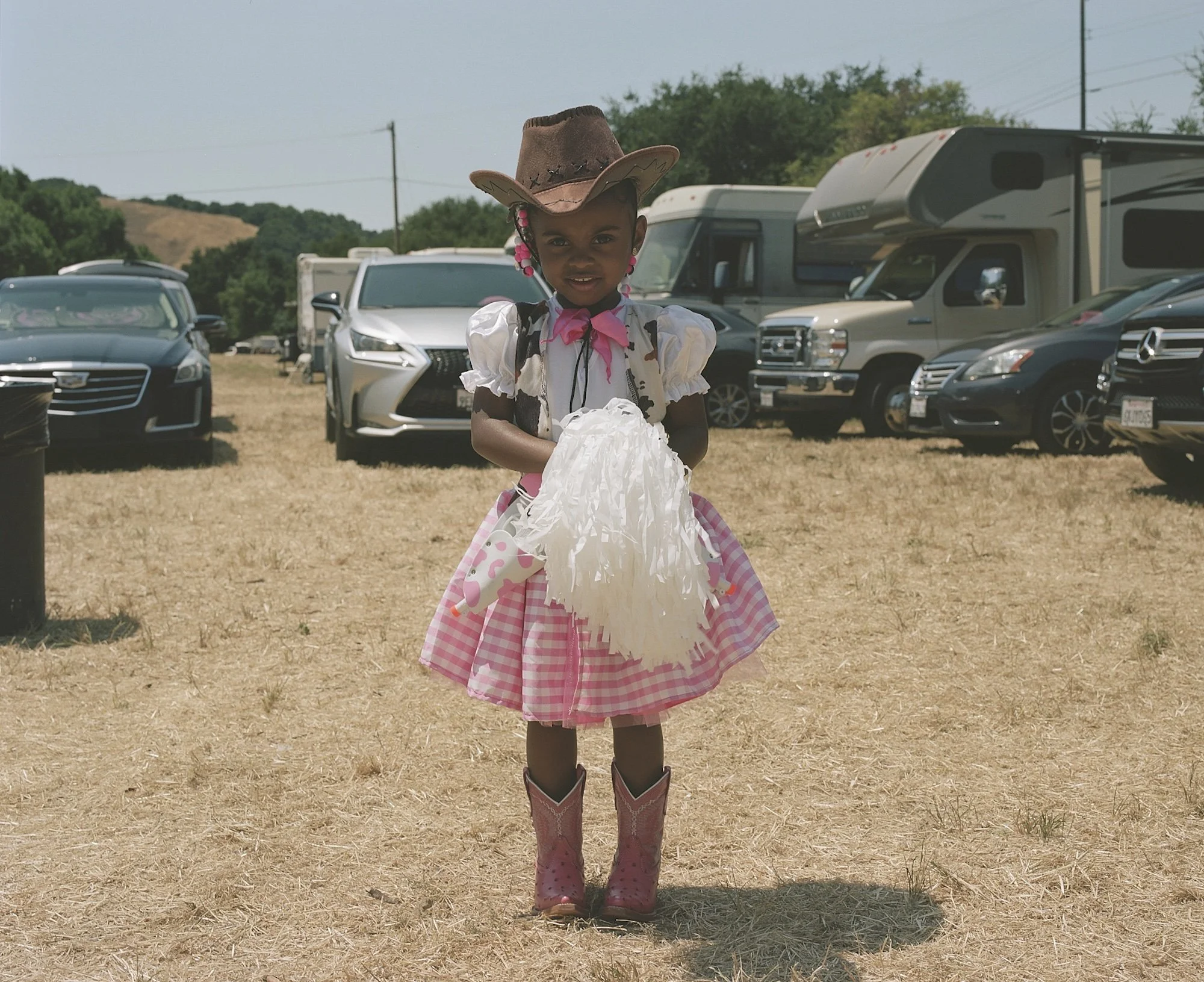 Young girl dressed as a cowgirl, wearing a pink checkered skirt, pink cowboy boots, and a cowboy hat, standing on dry grass in a parking lot with parked cars and RVs in the background.
