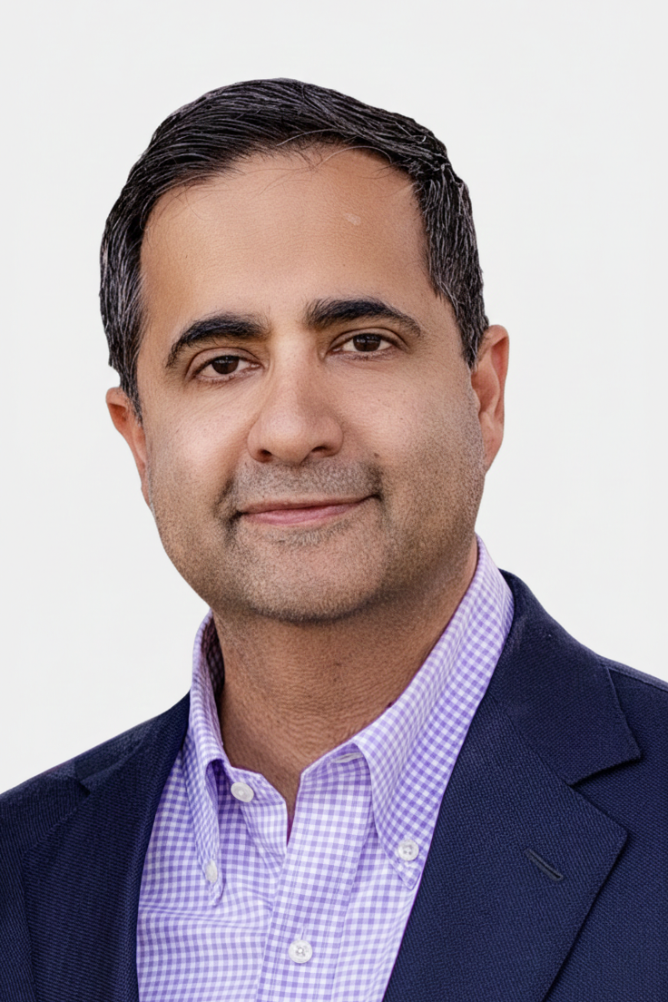 Portrait of a man with dark hair, wearing a purple checkered shirt and a navy blazer, against a plain white background.