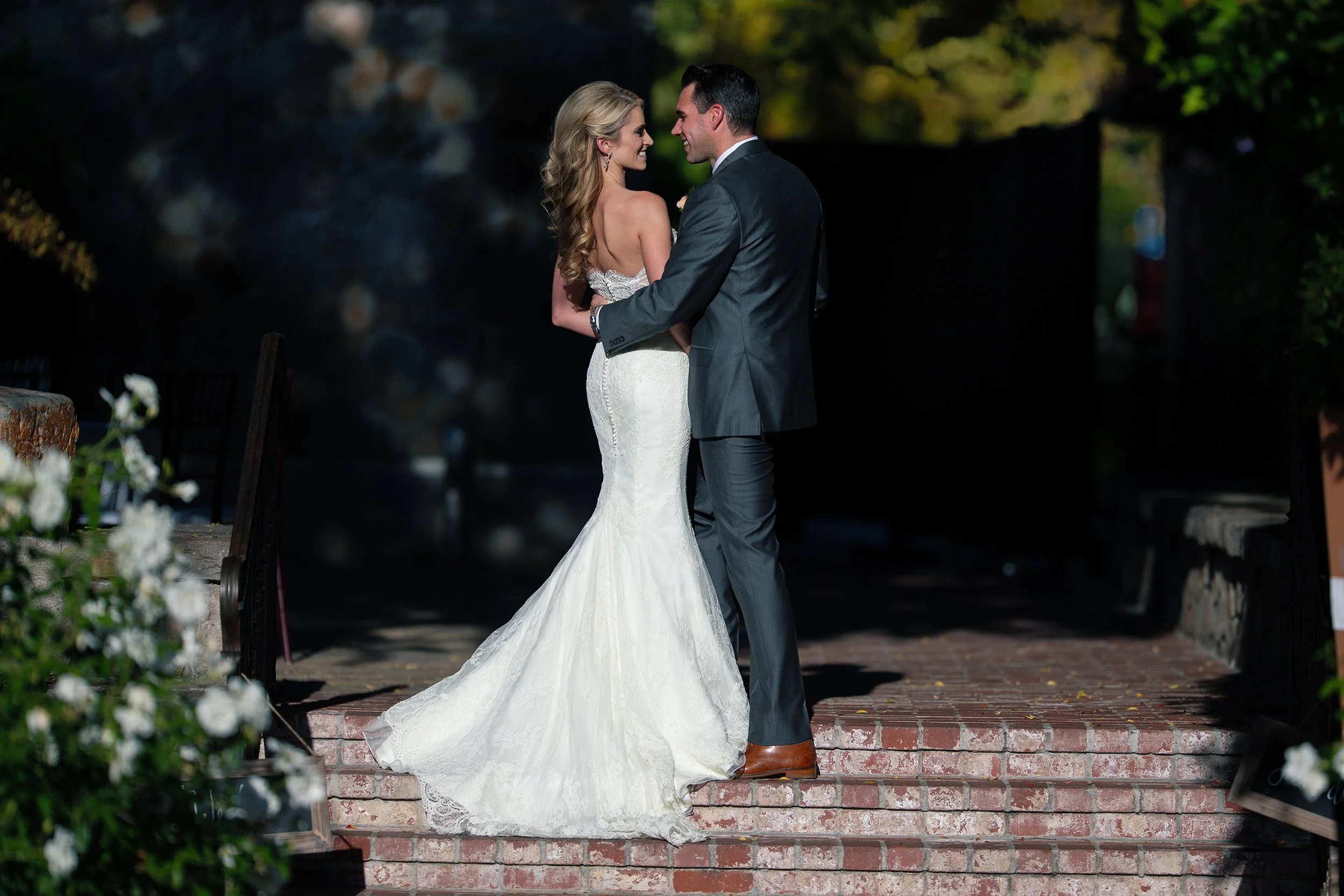 A bride and groom standing close together on brick steps outdoors, gazing into each other's eyes during their wedding ceremony, with trees and sunlight in the background.