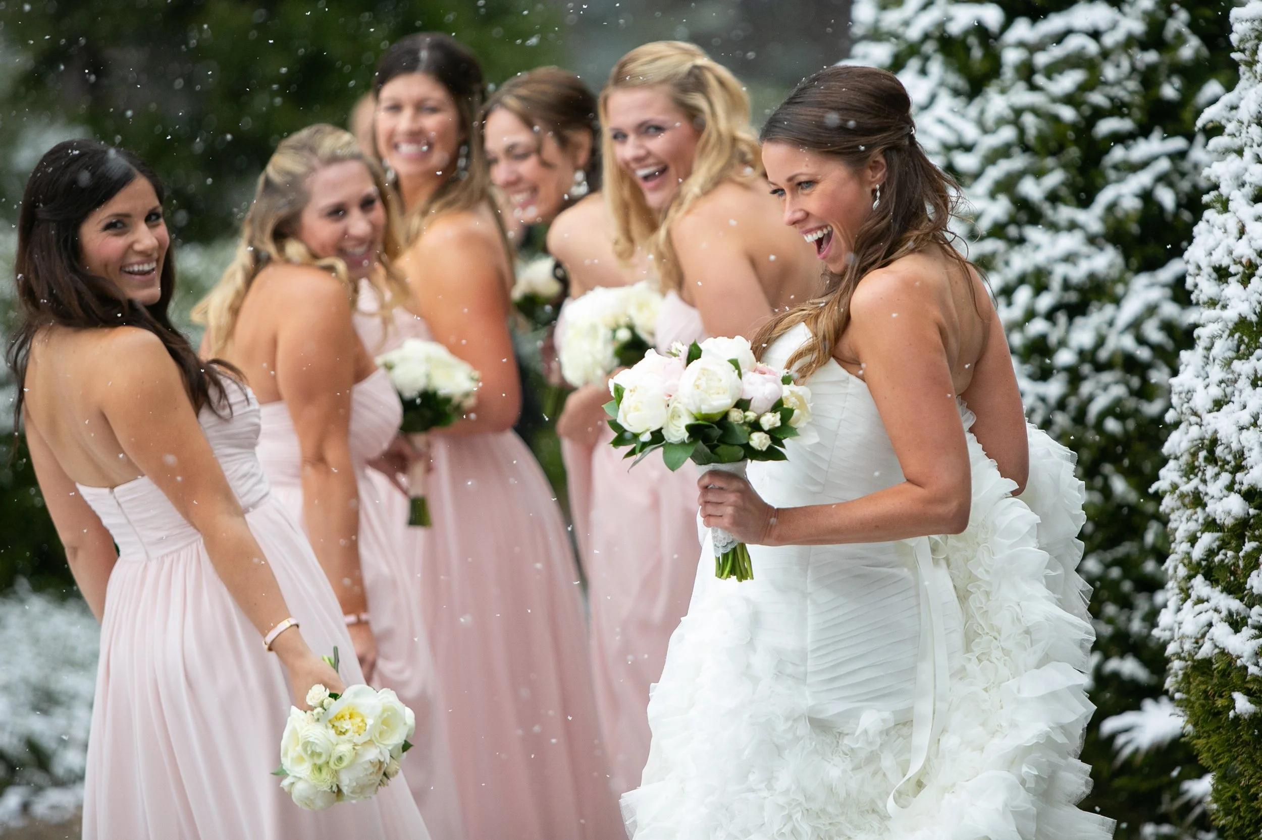 A bride and her bridesmaids outdoors in a snowy setting, all smiling and holding bouquets of white flowers. The bride is in a white wedding dress, and the bridesmaids are in light pink dresses.