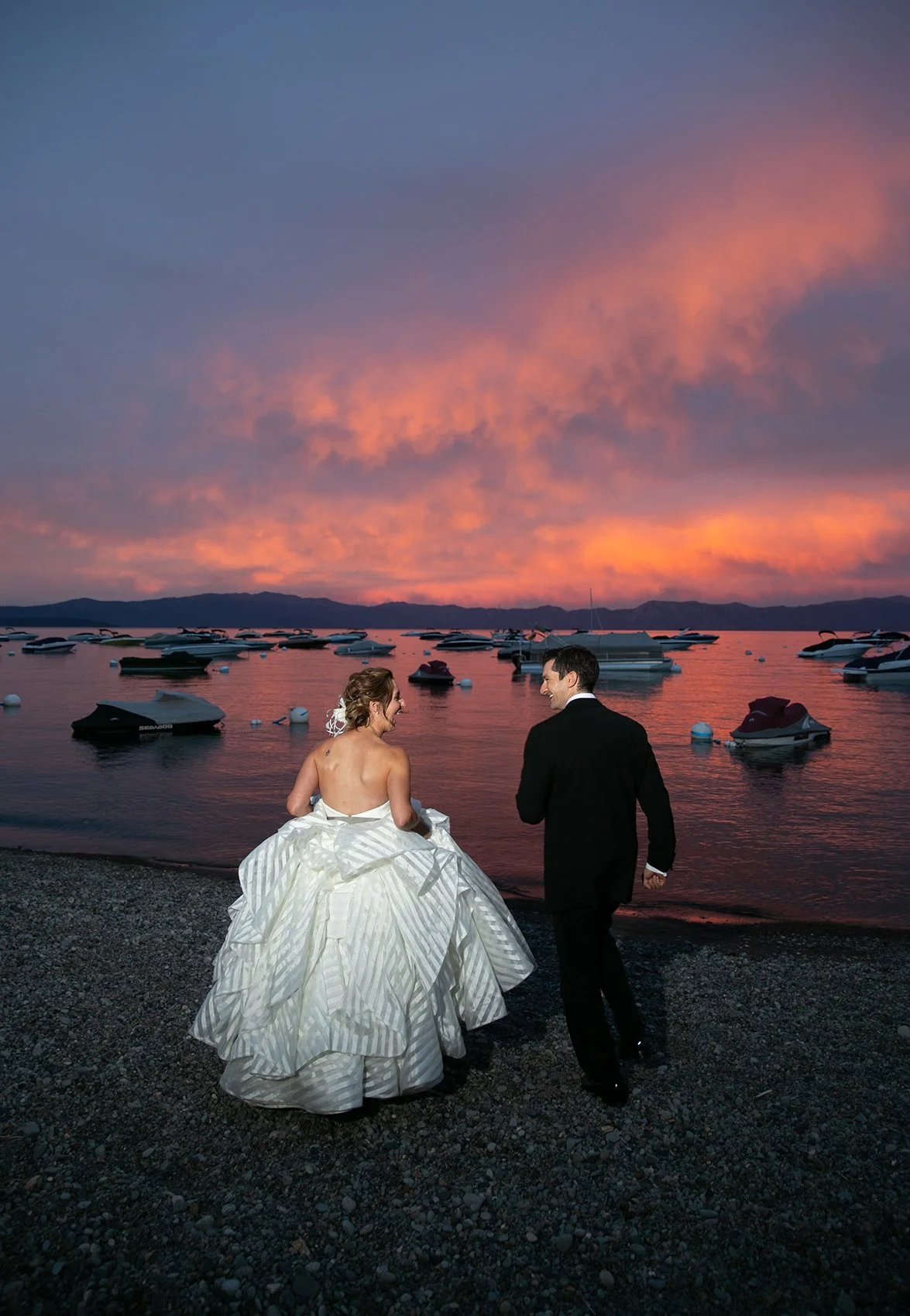 A bride and groom walking along a rocky beach at sunset, with boats anchored in the water and mountains in the background.
