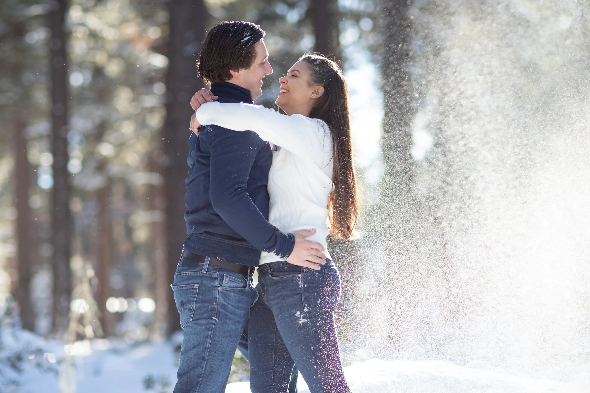 A young couple is embracing and smiling in a snowy forest, with sunlight shining through trees; the woman is wearing a white sweater and the man is in a dark jacket.