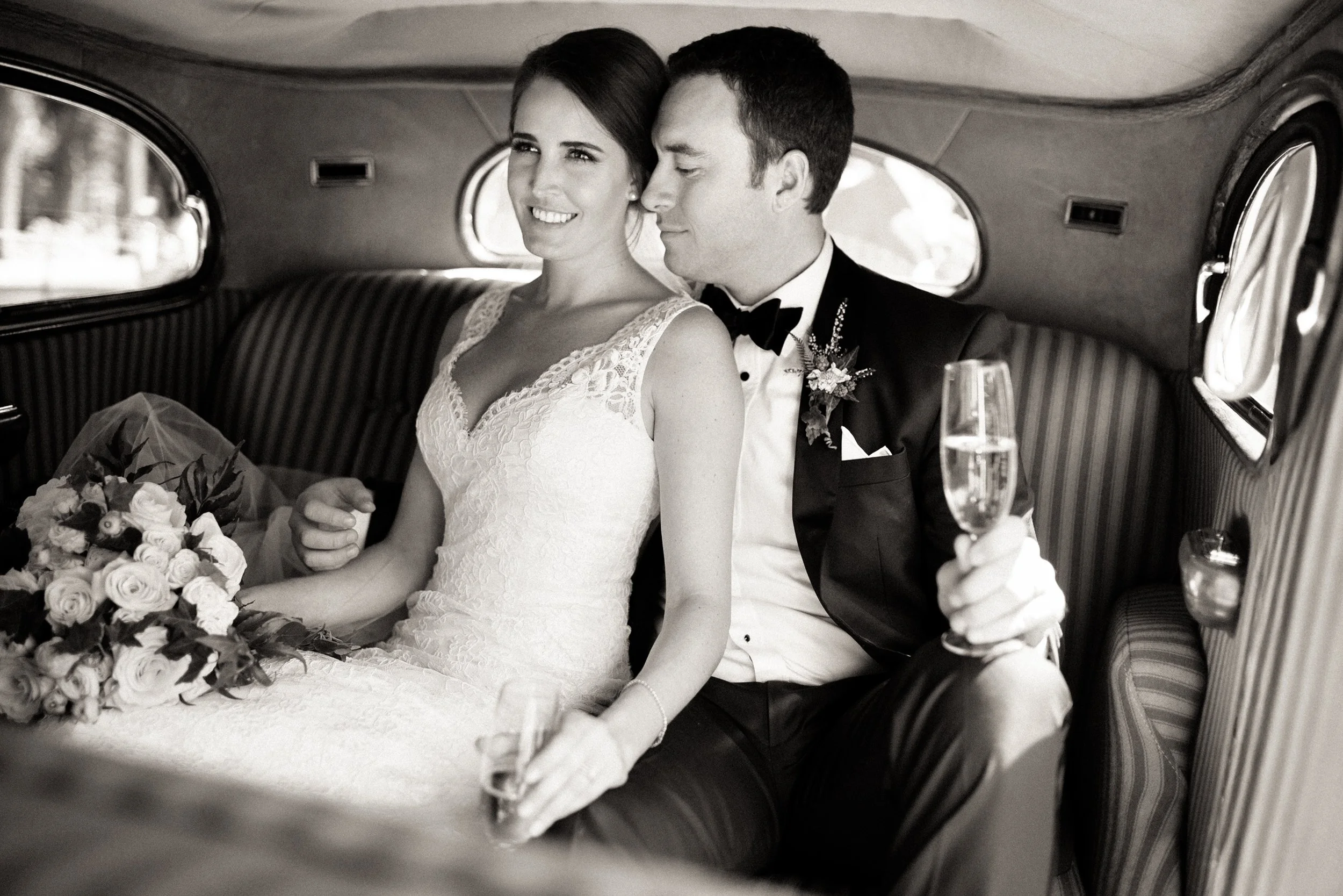 A bride and groom sitting inside a vintage car, celebrating their wedding. The bride is holding a bouquet, and the groom is holding a glass of champagne. The bride is smiling and looking out the window, while the groom is sitting close to her.