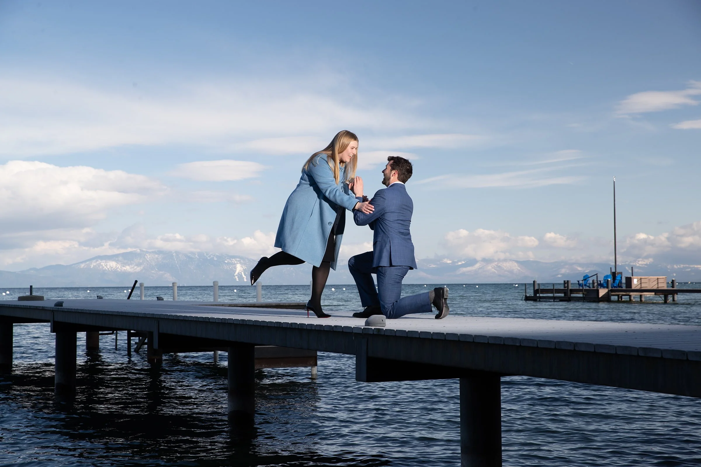A man in a suit kneeling on a dock, proposing to a woman in a coat, with a body of water and mountains in the background.