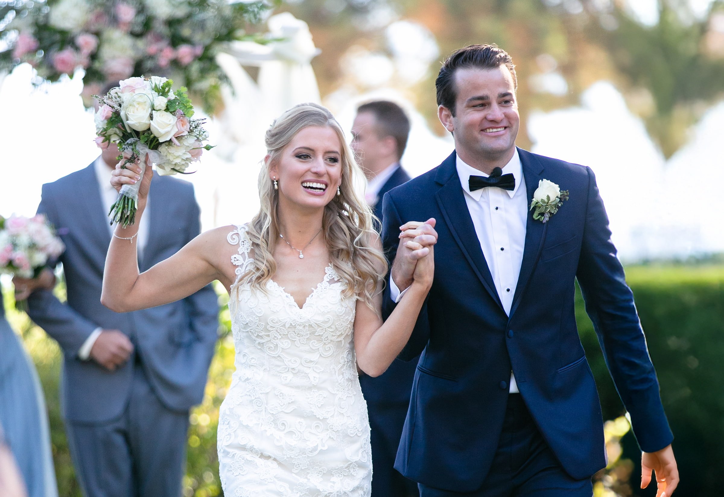 A smiling bride and groom walking outdoors, holding hands, with the bride holding a bouquet of flowers. The bride is wearing a white lace wedding dress, and the groom is in a navy blue tuxedo with a black bow tie. In the background, there are guests and greenery.