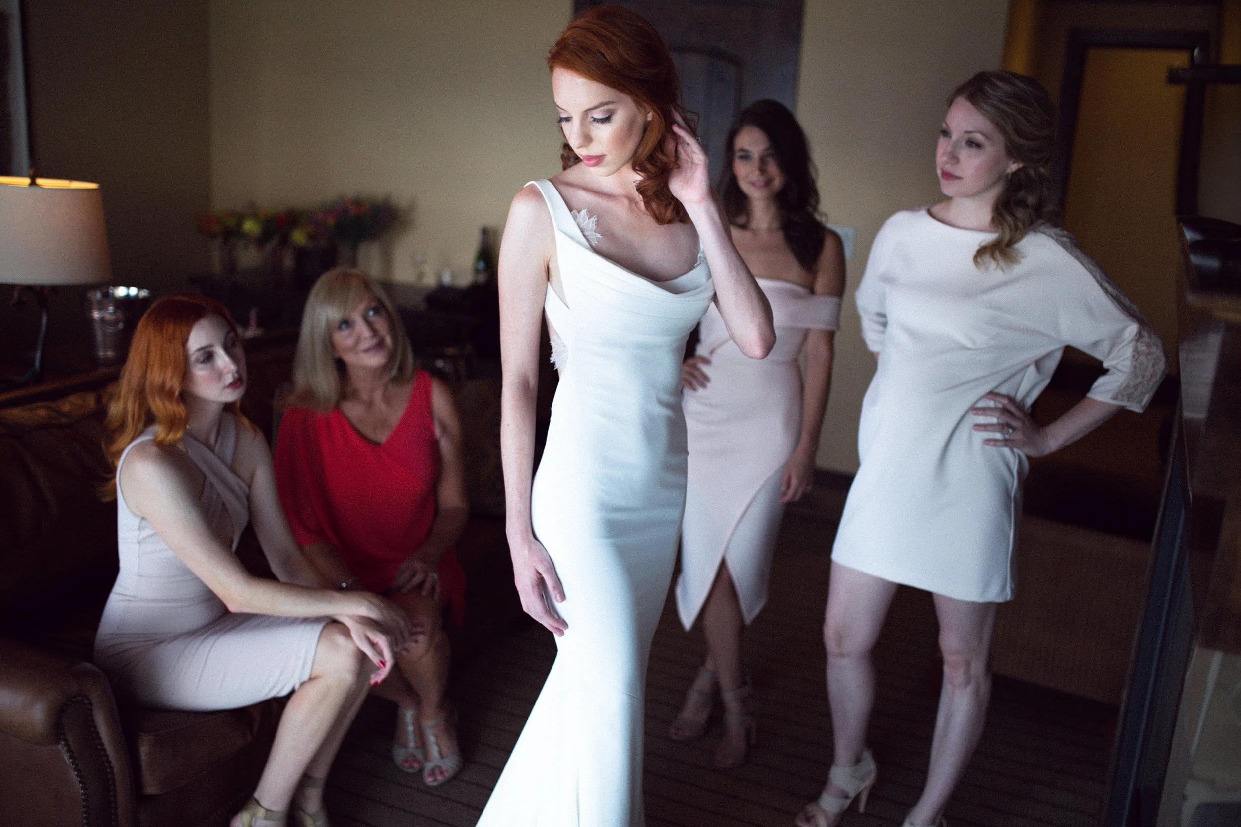 Five women, one in a white wedding dress, are in a room with a brown leather couch. The bride is standing and looking down, while the other four women sit or stand around her, observing her.