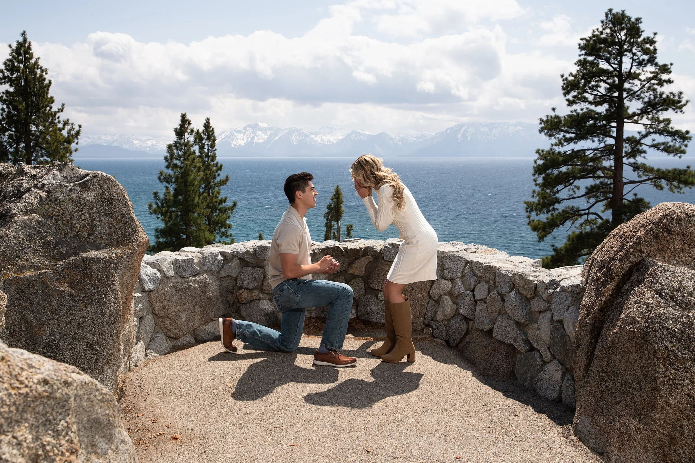 A man proposing to a woman near a stone railing overlooking a lake and snow-capped mountains, with pine trees and partly cloudy sky in the background.