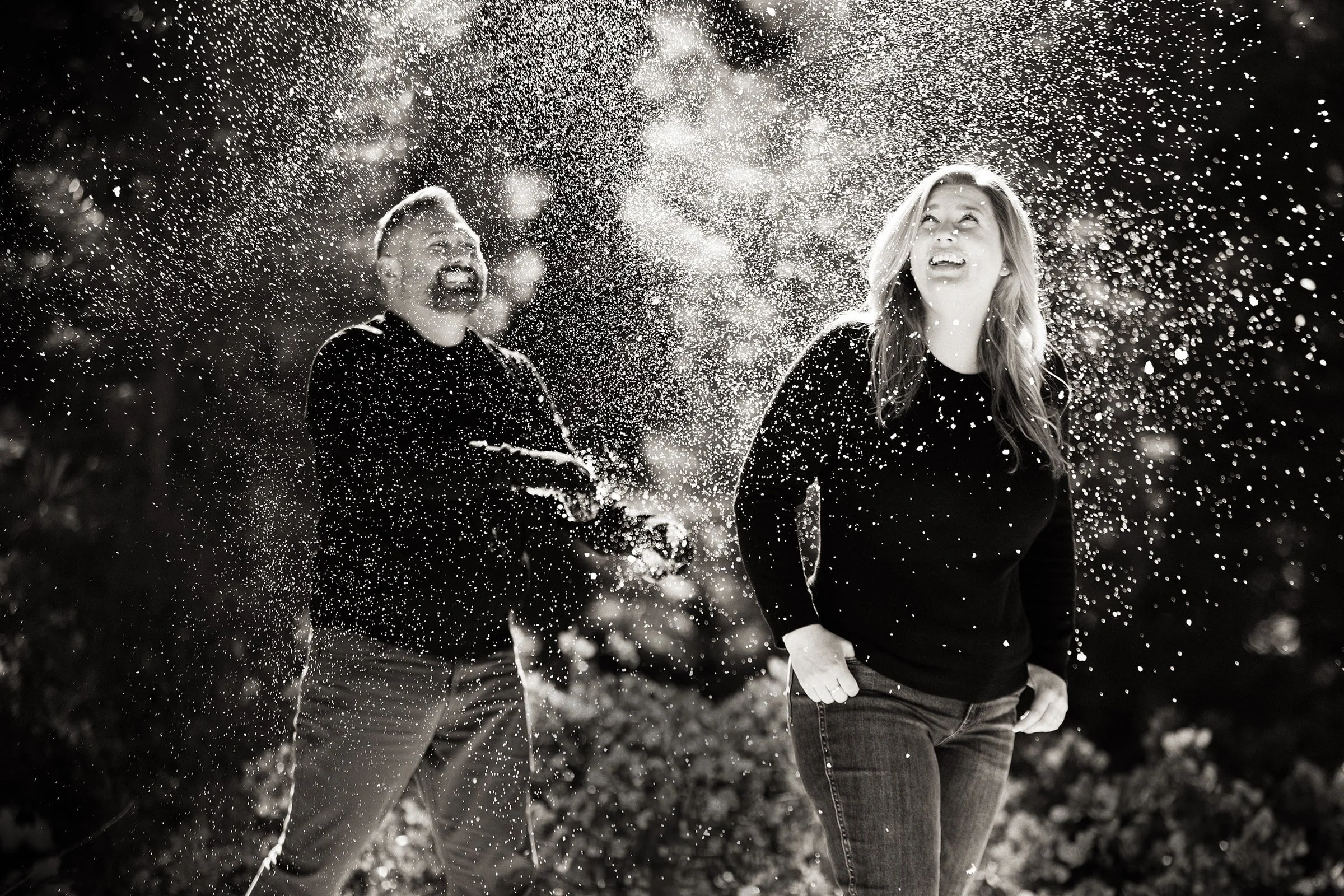 A man and a woman laughing and playing with water outdoors at night, with water splashing around them in a joyful scene.