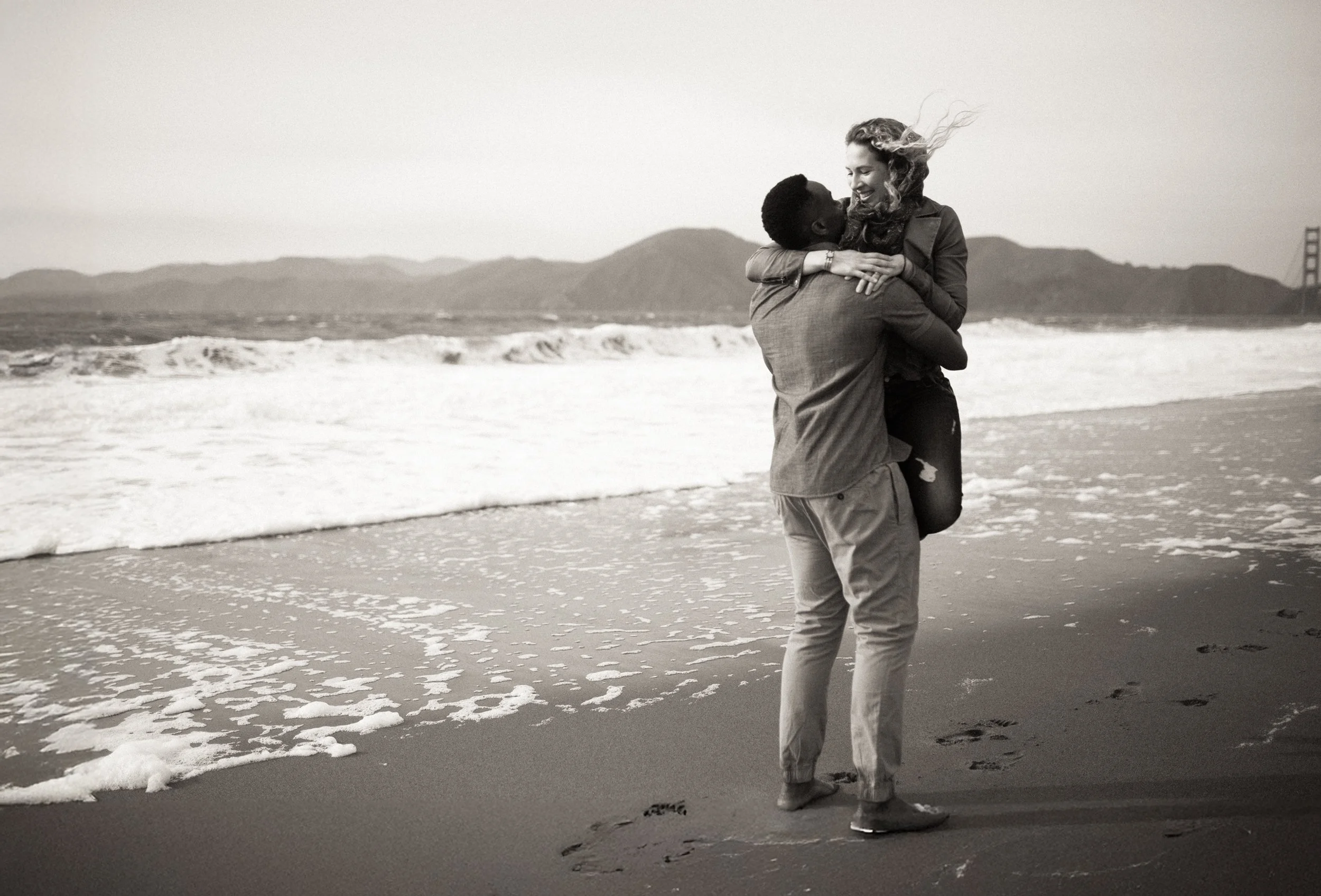 A couple embracing on a beach, with the man lifting the woman off the ground, both smiling and looking at each other, with waves and mountains in the background.