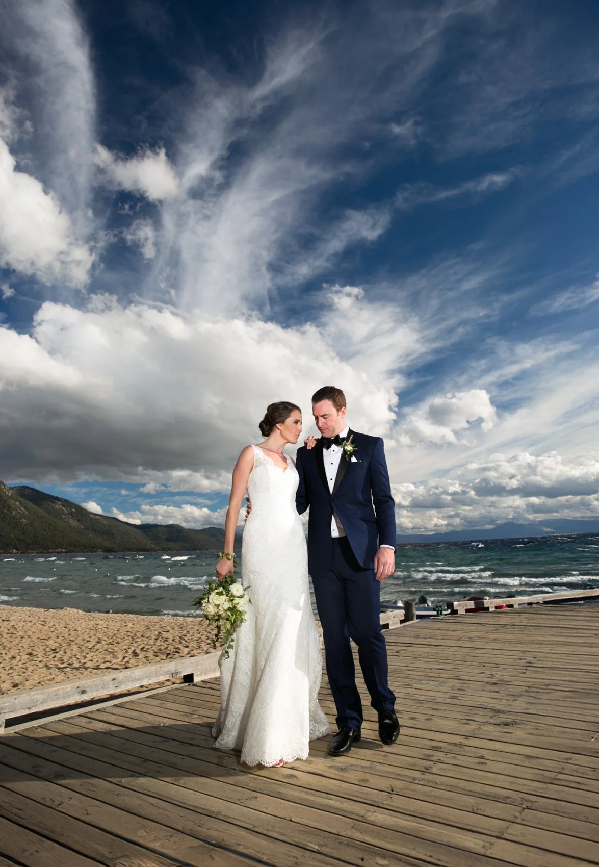 A bride and groom standing on a wooden dock by the water on their wedding day, with a cloudy sky and mountains in the background.