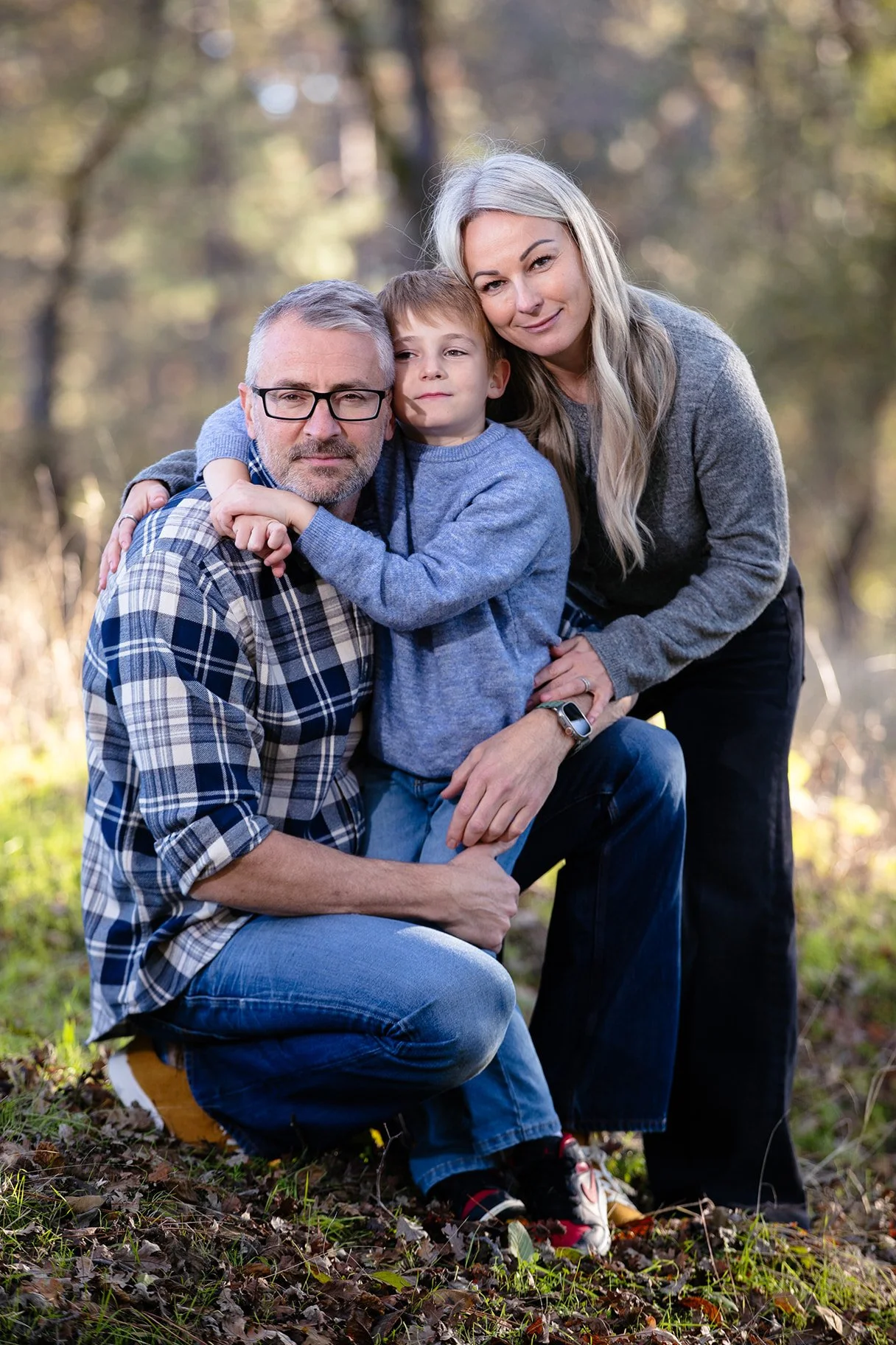 Family of three outdoors in a wooded area, embracing and looking at the camera.
