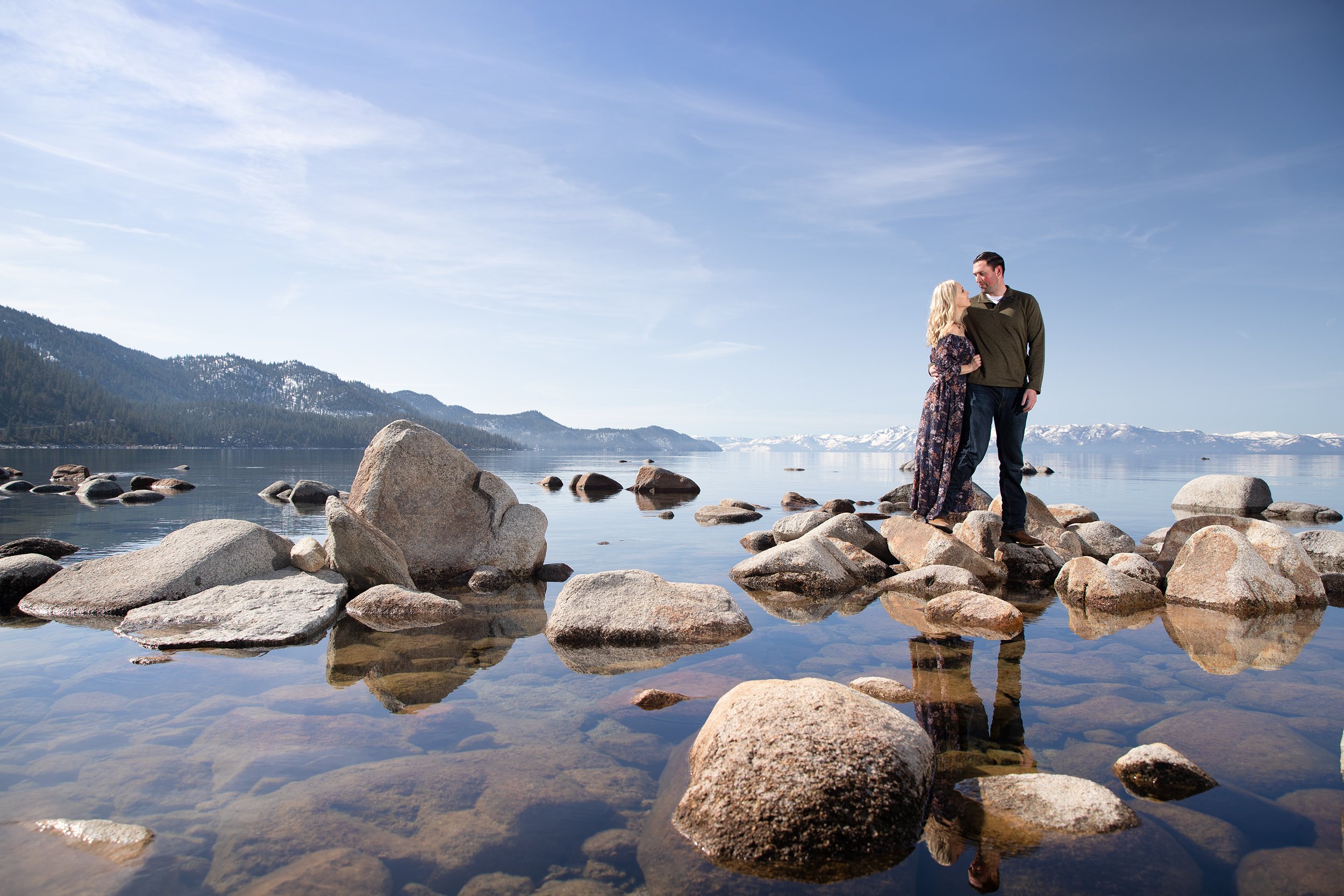 A couple standing on rocks in a calm lake, with mountains and snow in the distance under a blue sky