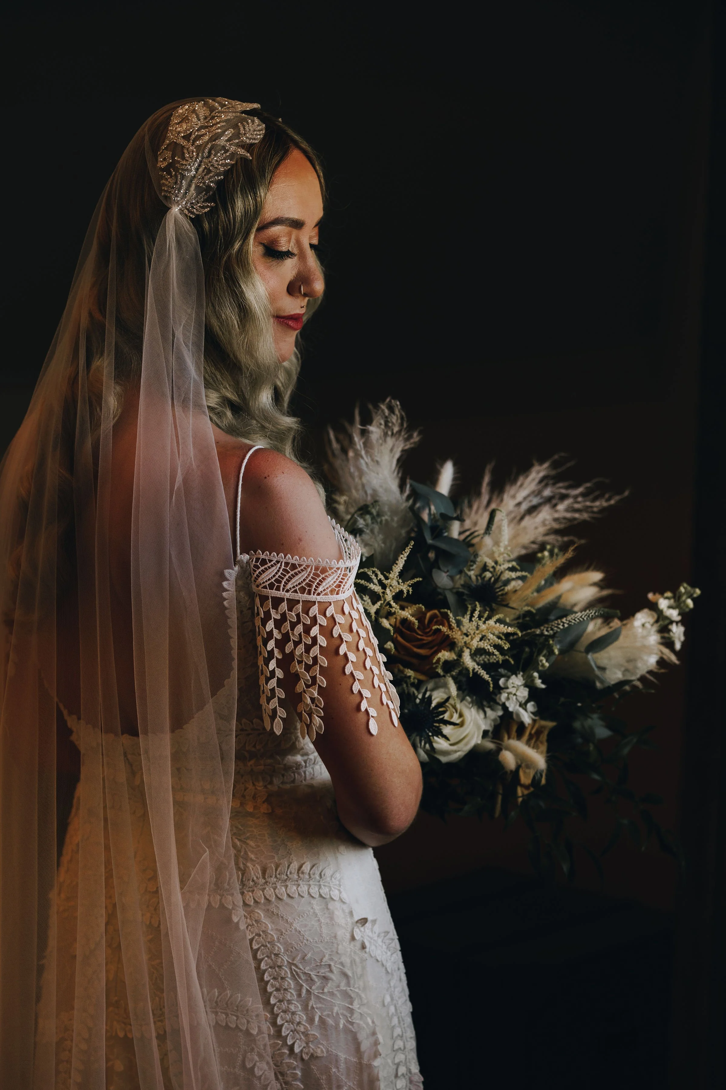 A bride in a embroidered wedding dress with a veil, holding a bouquet of flowers, standing in front of a dark background.