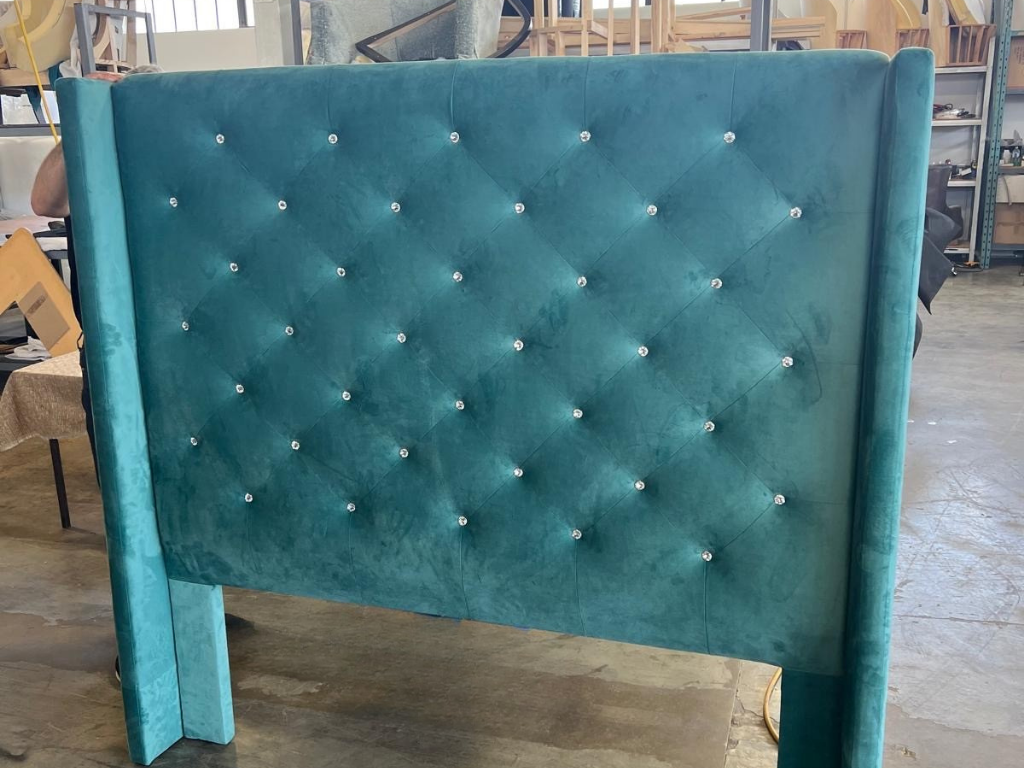 Teal upholstered headboard with diamond-tufted pattern and rhinestone accents, standing vertically in a workshop or store.