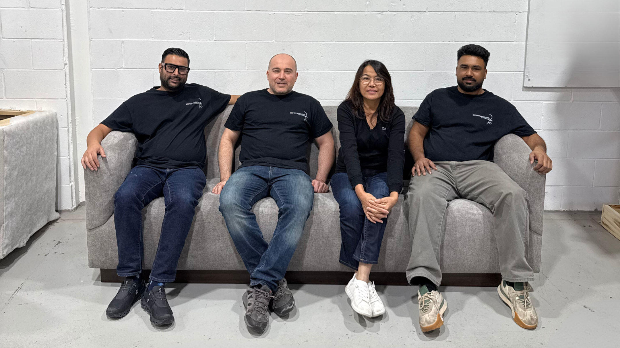 Four people sitting on a gray couch against a white brick wall, all wearing black T-shirts, with three men and one woman.