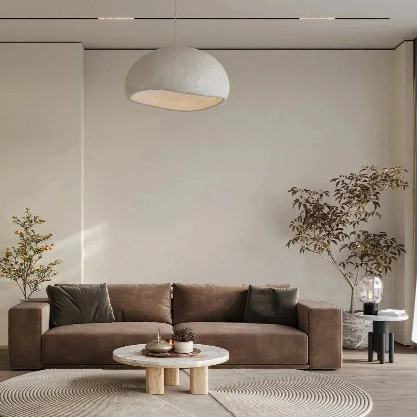 A living room with a beige sofa, two dark pillows, a small table with decorative items, a large potted plant, and a hanging ceiling light, with minimalist decor and neutral tones.
