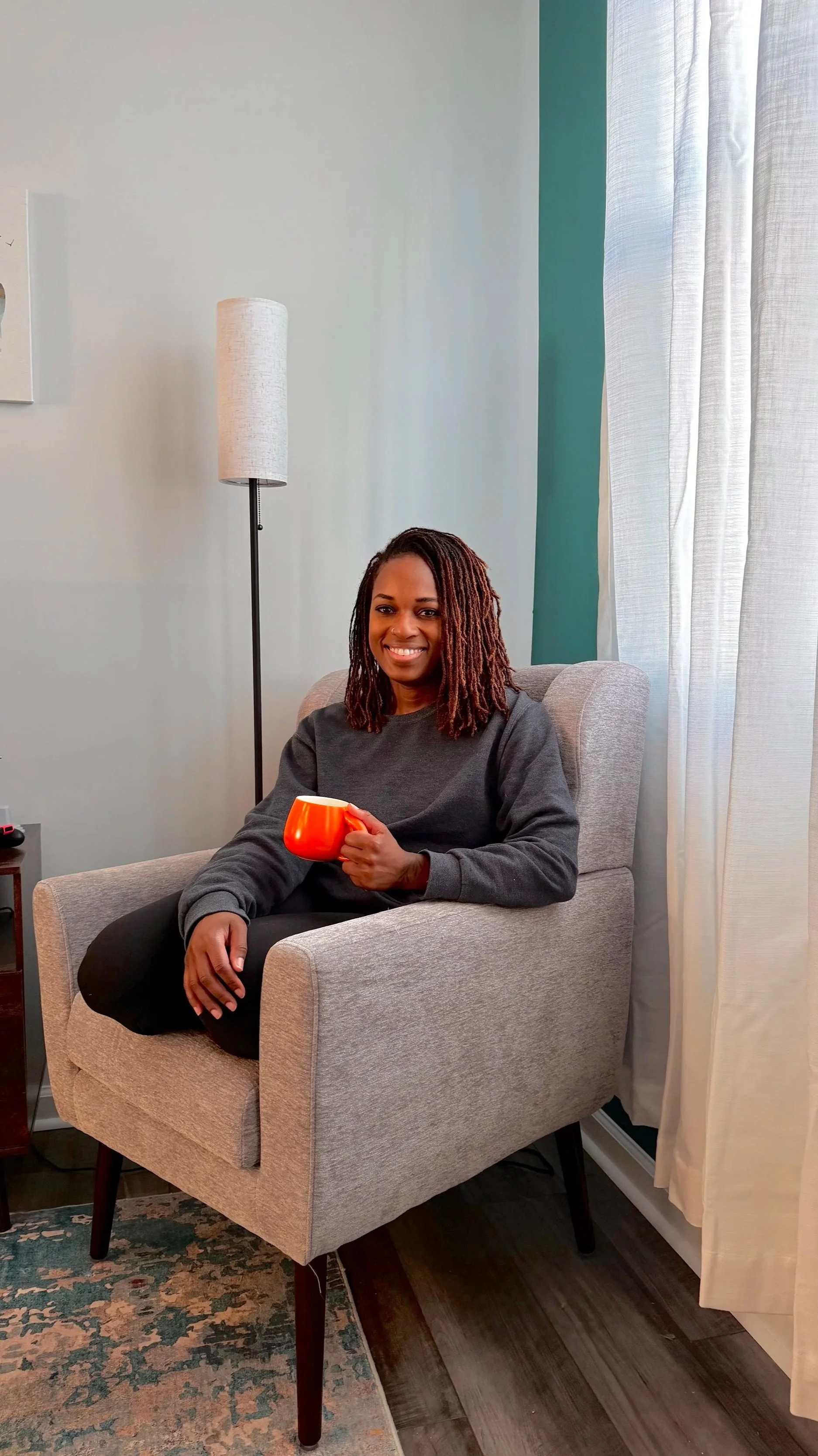 A woman sitting in a beige armchair, holding a red mug, smiling, with a lamp, curtain, and wall in the background.