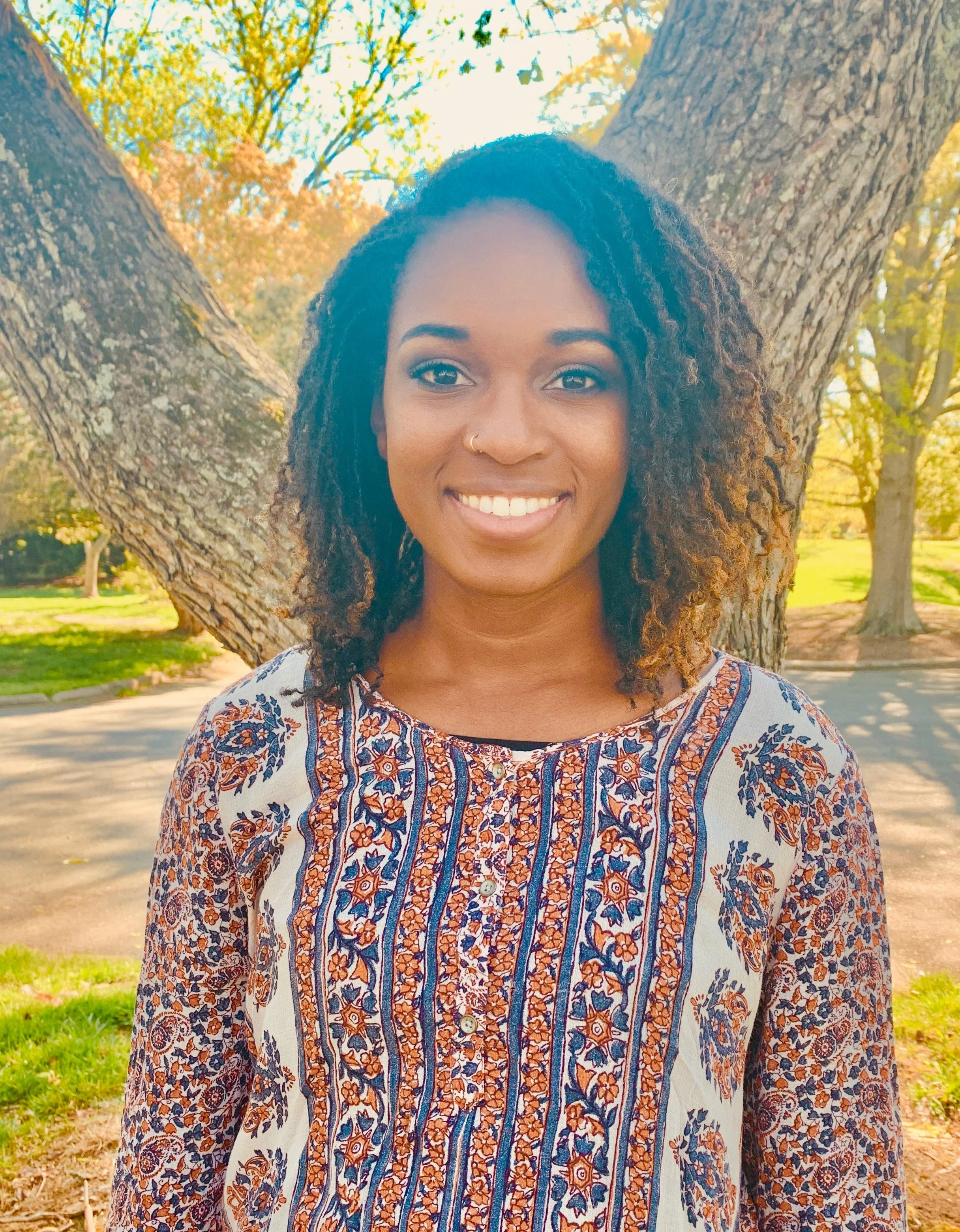 A woman with curly hair and a nose piercing smiling outdoors in front of a tree with autumn leaves.
