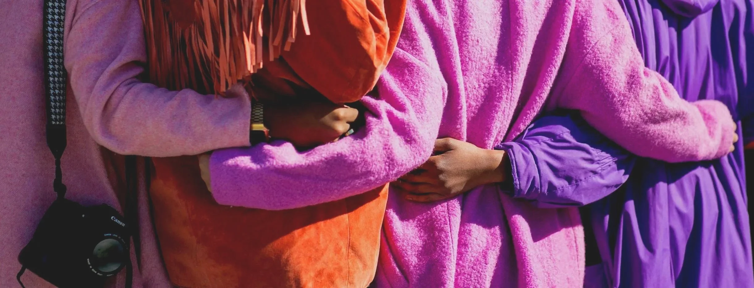 Close-up of several people standing in a line, wearing colorful warm clothing, with arms crossed or holding hands, and a camera hanging from a strap.