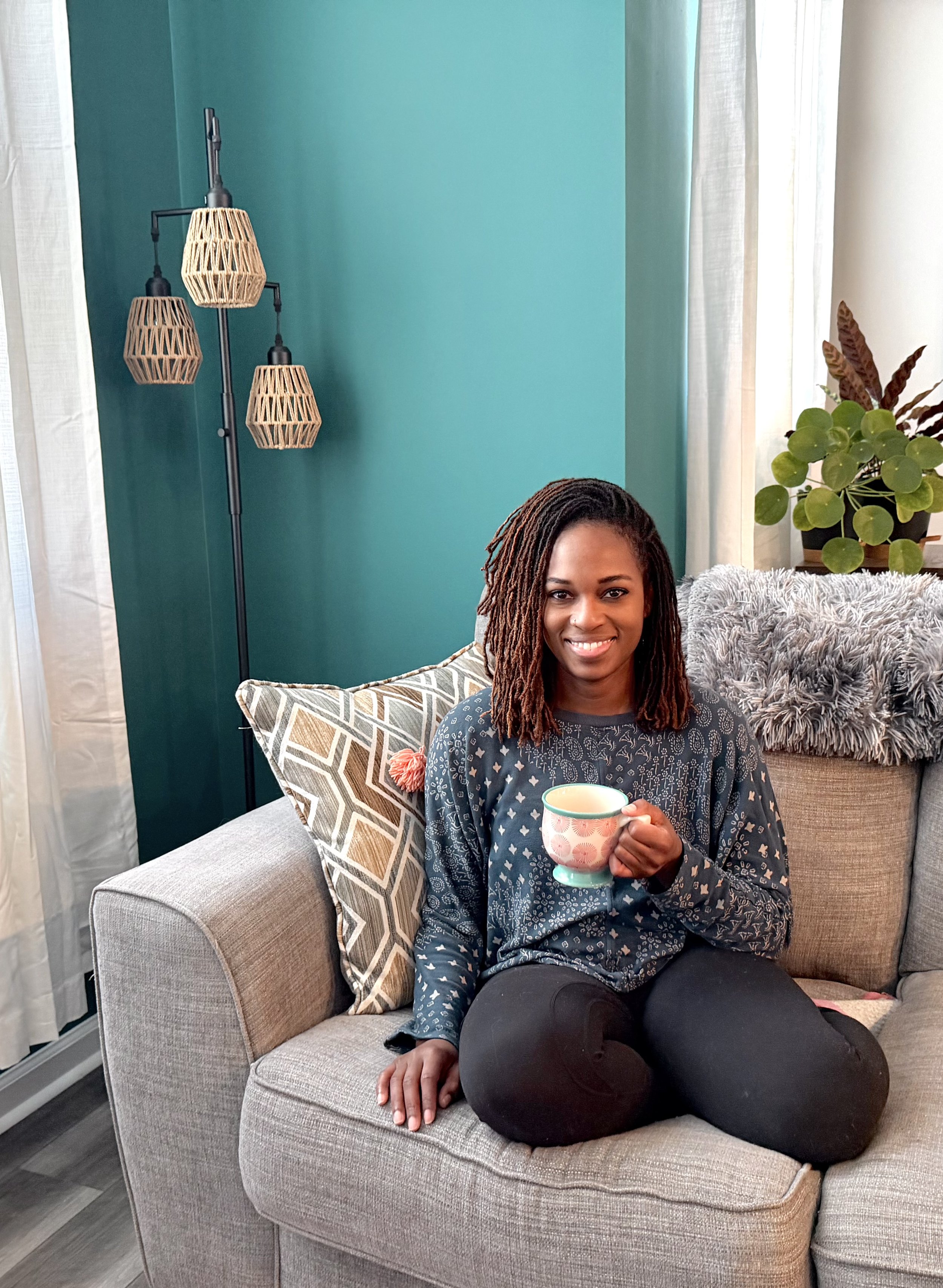 A woman with dark skin and curly shoulder-length hair, sitting cross-legged on a beige couch, holding a mug, smiling at the camera with a teal wall and decorative pillows behind her.