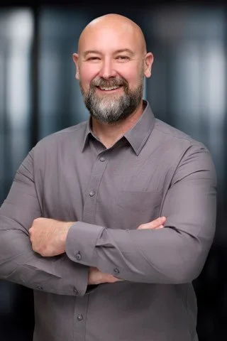 Gabe Smith, a smiling man with a beard and bald head, wearing a gray shirt, standing with arms crossed in front of a blurred office background.