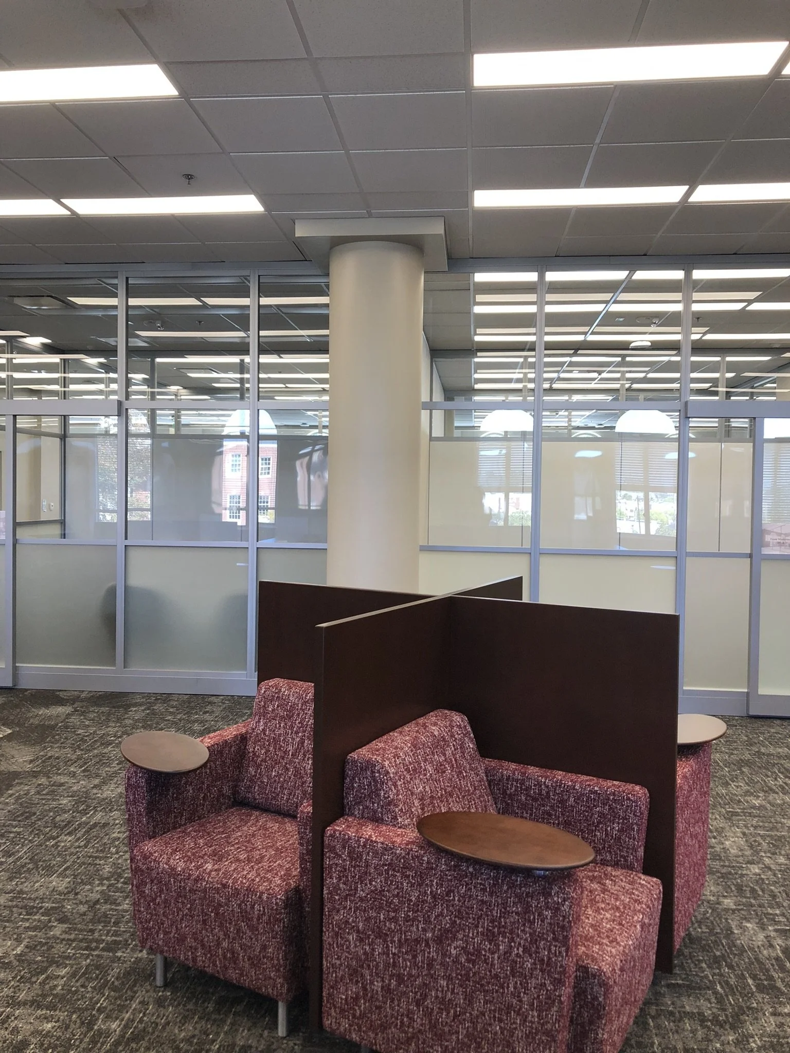 An interior view of library seating with red upholstered chairs equipped with small wooden tablet arms, arranged around a dark wood partition, with glass walls and ceiling lights visible in the background - project by B Group Architecture, Inc
