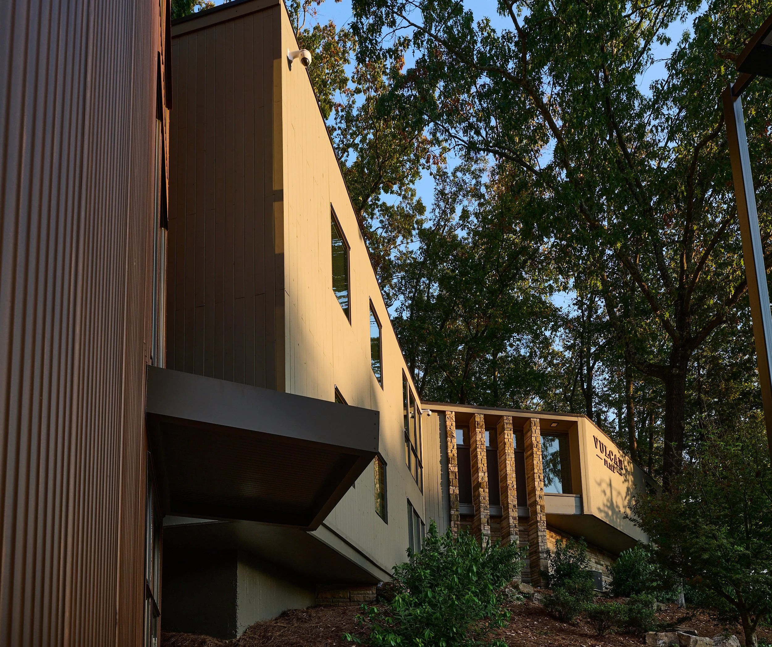 Modern multi-story building with large windows, surrounded by trees and greenery, with sunlight casting shadows.