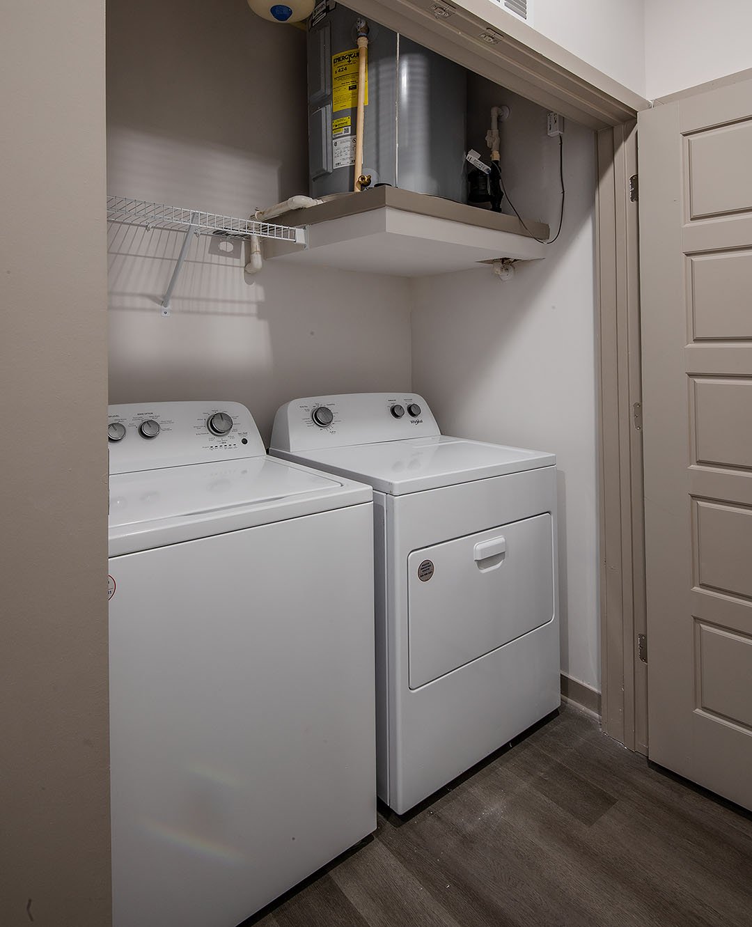Laundry room with white top-loading washing machine and front-loading dryer, a wire shelf above, and a utility area with a water heater and pipes.