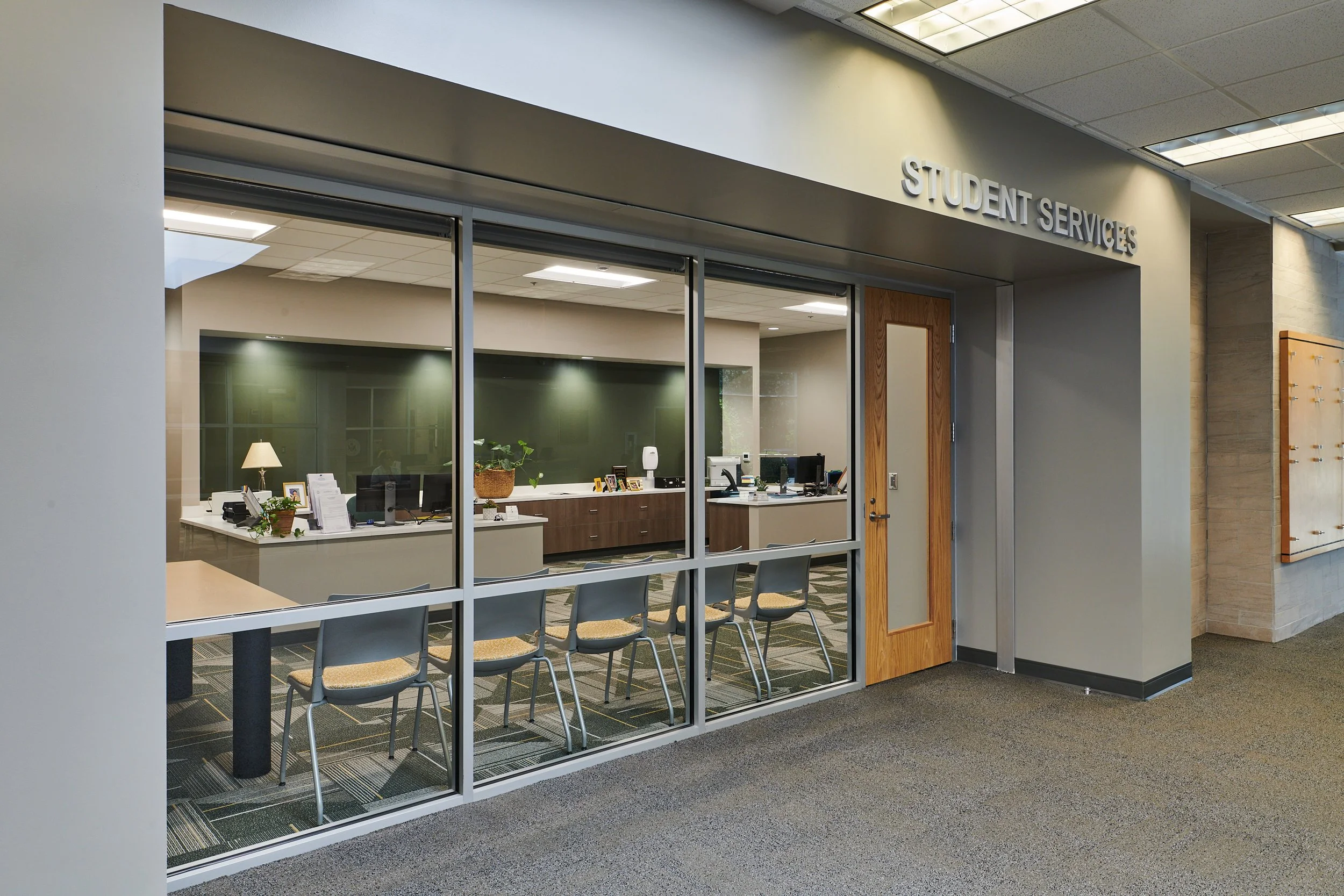 View of a student services office with glass walls, a wooden door, and a sign above that reads 'STUDENT SERVICES'. Inside, there are desks, chairs, and office supplies -project by B Group Architecture, Inc.