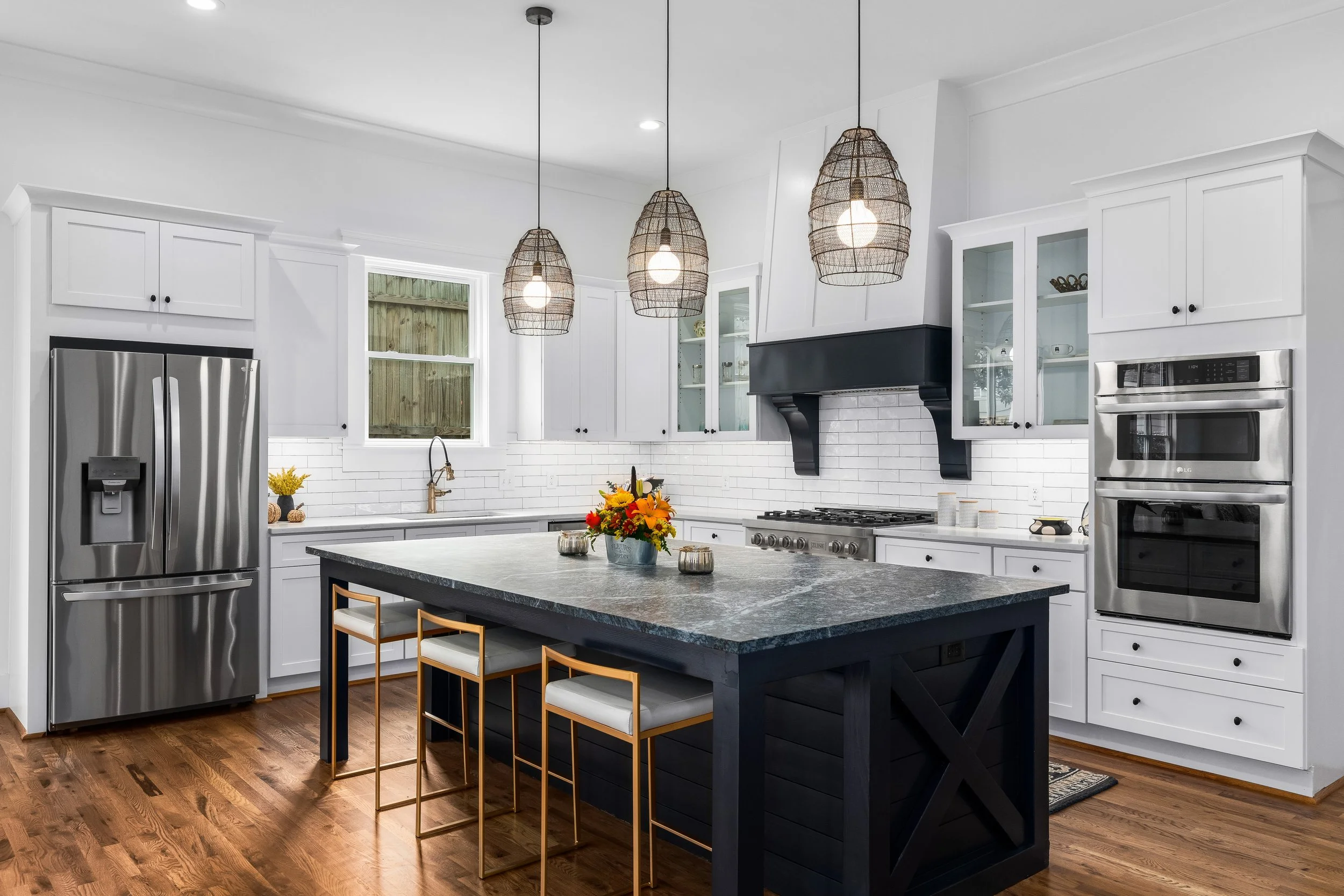 Modern kitchen with white cabinetry, stainless steel refrigerator and ovens, black island with a flower arrangement, hanging wicker pendant lights, wood flooring, and a white subway tile backsplash.