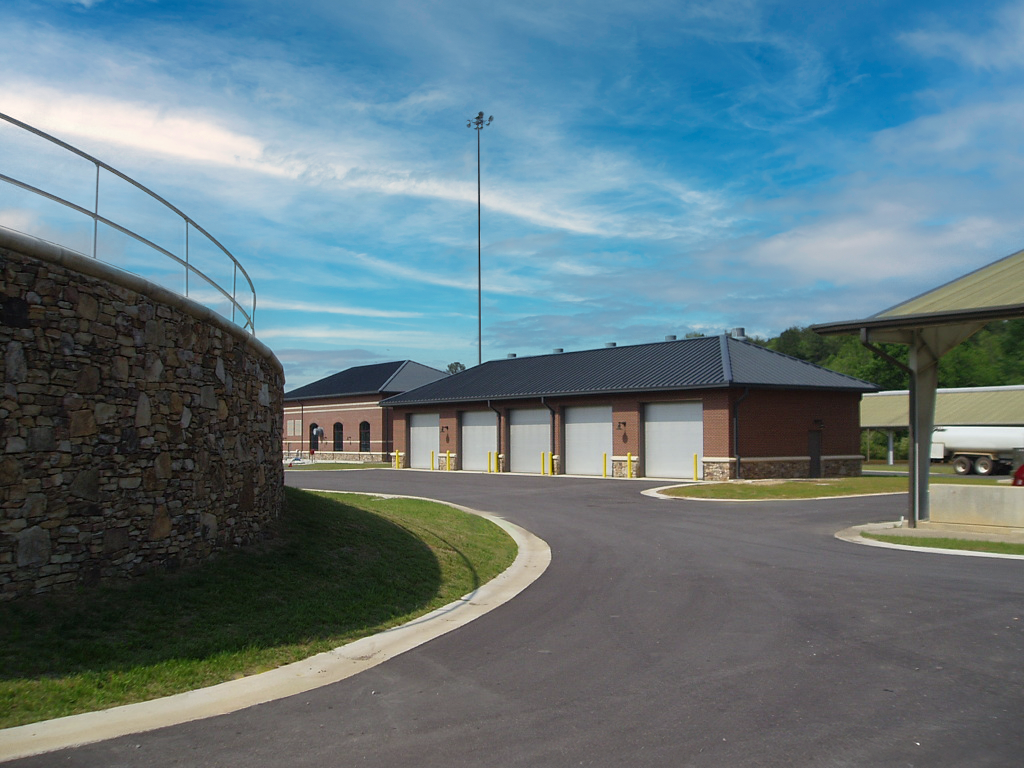 A paved driveway curves past a stone, leading to a building with multiple garage doors, adjacent to a covered parking area. 